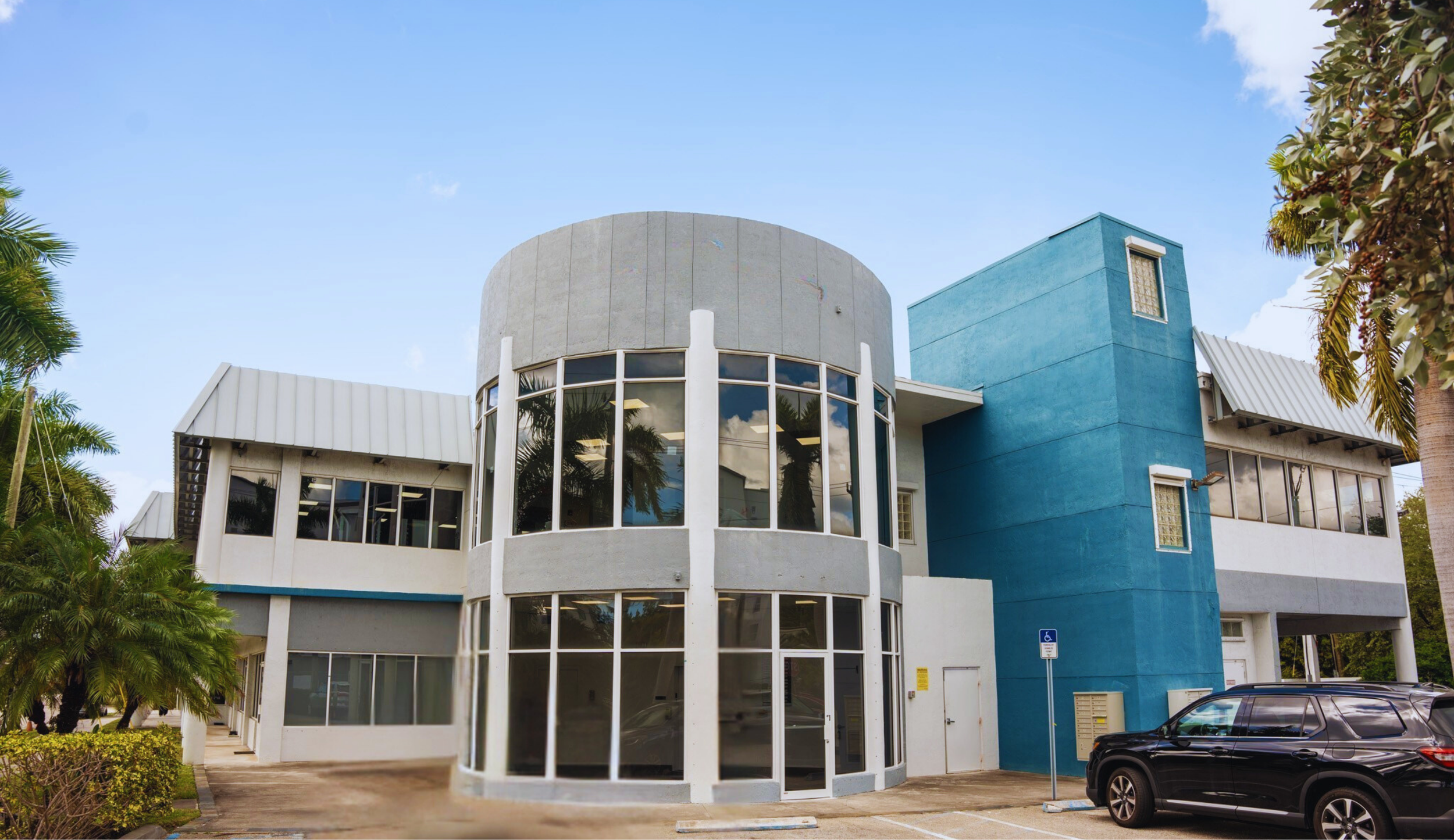 Modern two-story building with a curved glass front, blue and gray exterior walls, and surrounding palm trees, with a parking lot in the foreground.