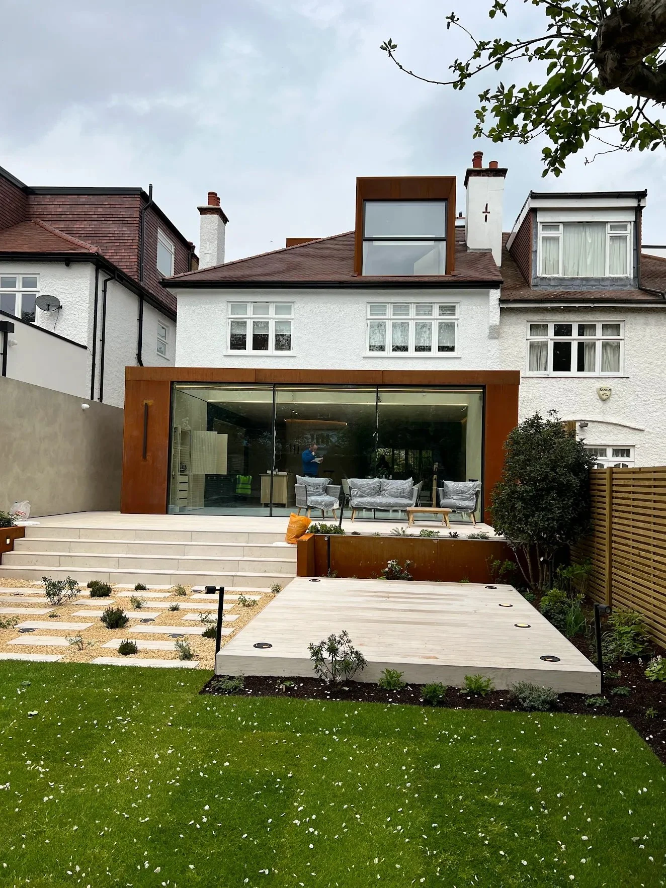 Modern house with a glass back wall, multiple windows, and a small front garden with grass, a wooden patio, and garden beds.