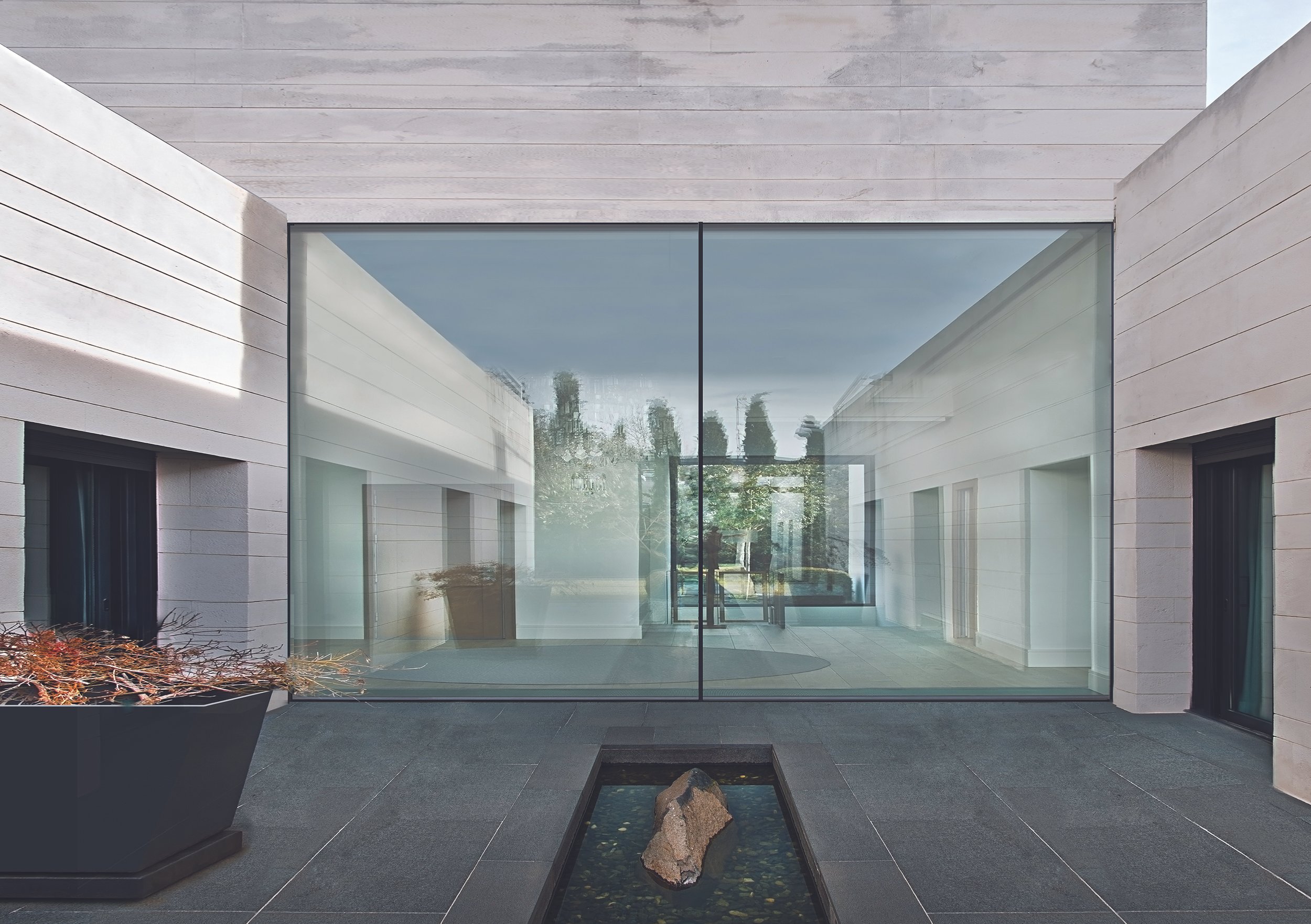 Modern building entrance with large glass doors, stone walls, dark gray tiled flooring, potted plant, and a small water feature with a rock in the foreground.