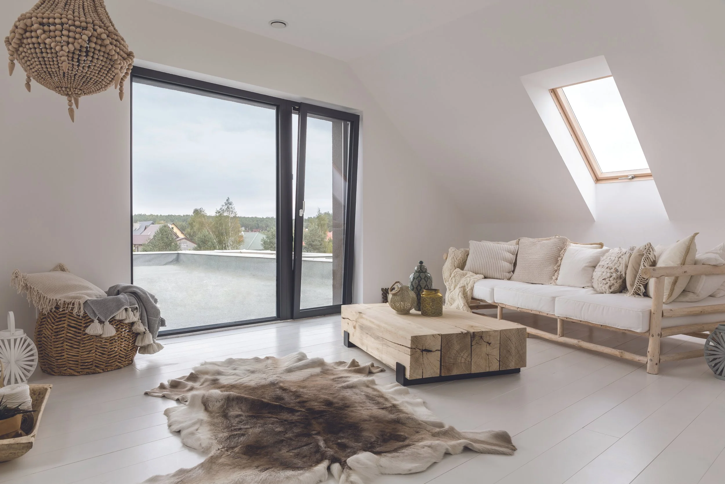 Bright living room with large sliding glass door and skylight, featuring a white sofa with neutral pillows, a rustic wooden coffee table, animal hide rug, woven basket, and decorative lanterns.