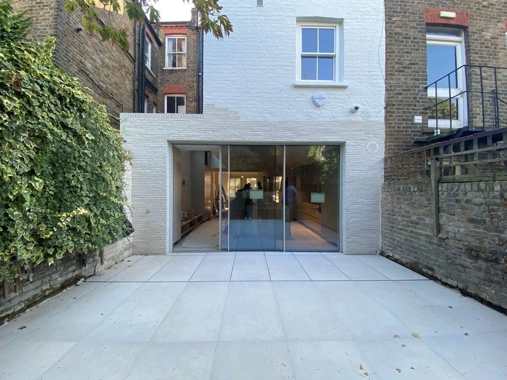 A modern house with a white exterior wall and large glass sliding doors, surrounded by a concrete patio and flanked by brick walls and greenery.