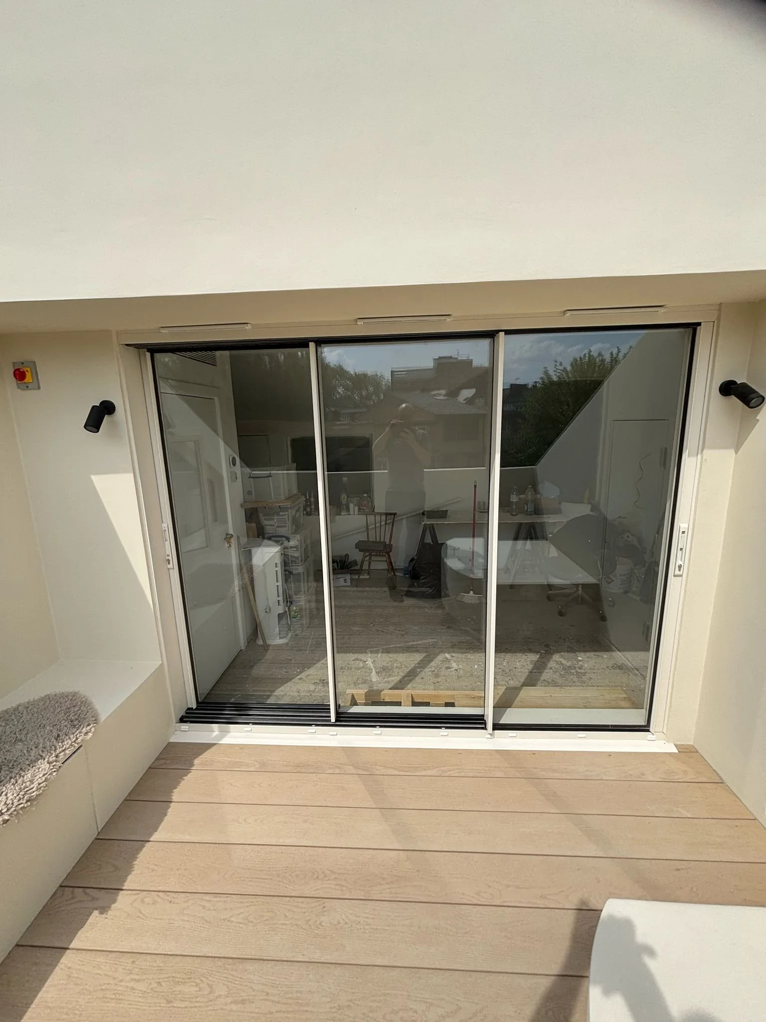 View of a sliding glass door opening to a small balcony with light wooden decking, view of an indoor cluttered room, and a reflection of the person taking the photo.