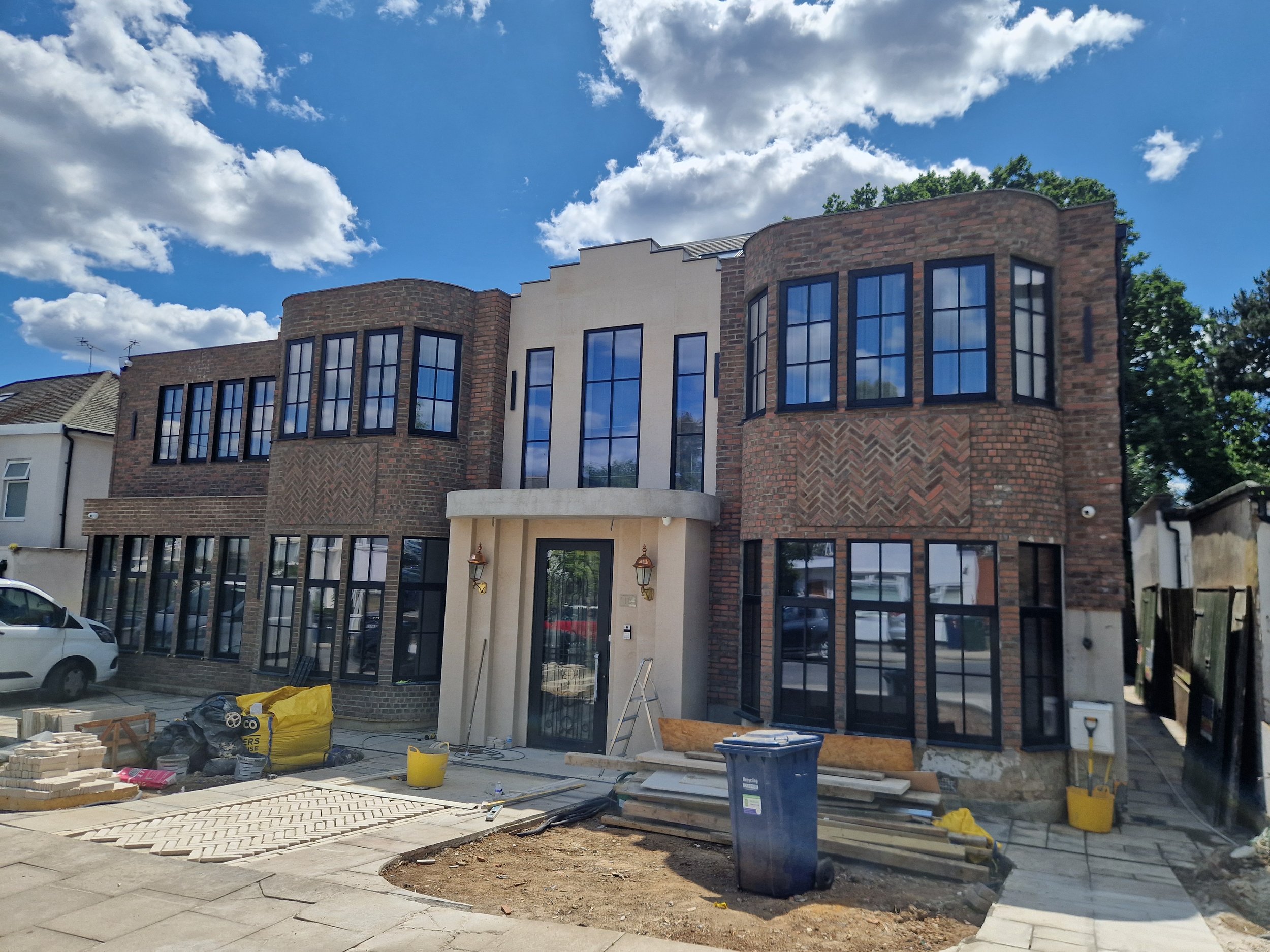 Construction of a modern multi-story brick house with large black-framed windows, street-level sidewalk, construction materials, and blue sky with clouds.