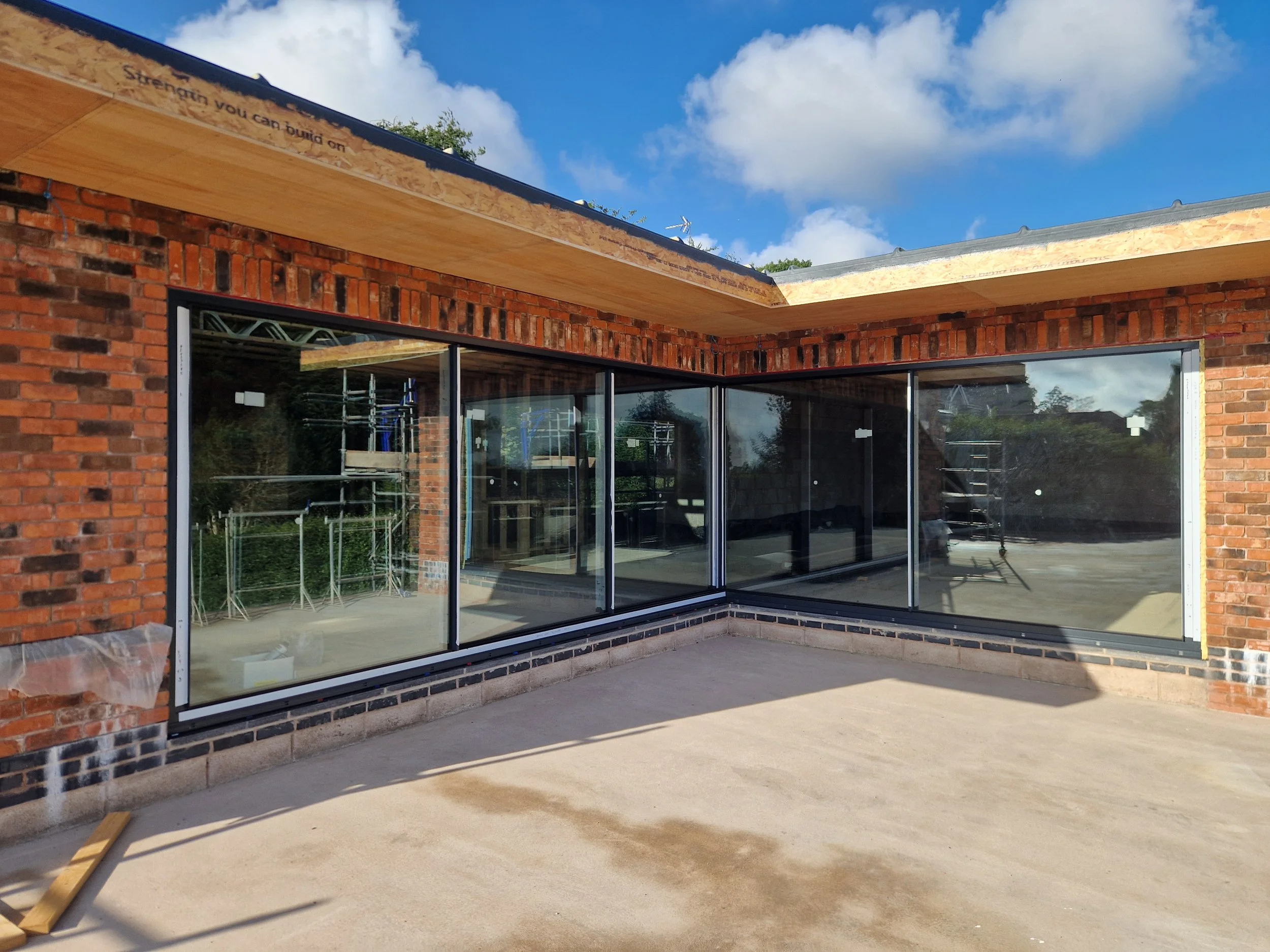 Construction site of a brick building with large glass windows and a partially built roof under a blue sky with clouds.