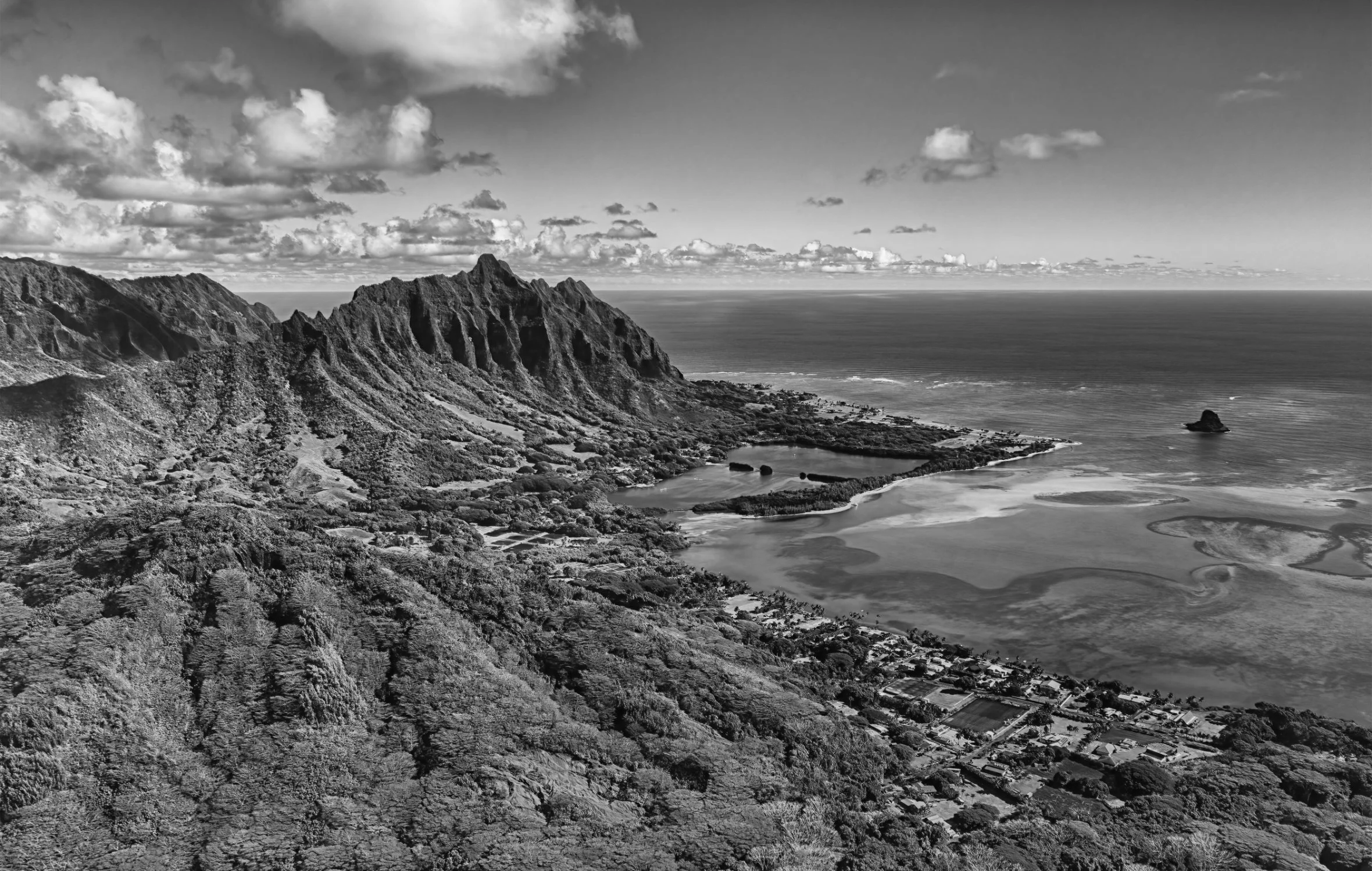 A black and white aerial view of a coastal landscape with mountains, a lagoon, a sandy shore, and small buildings.