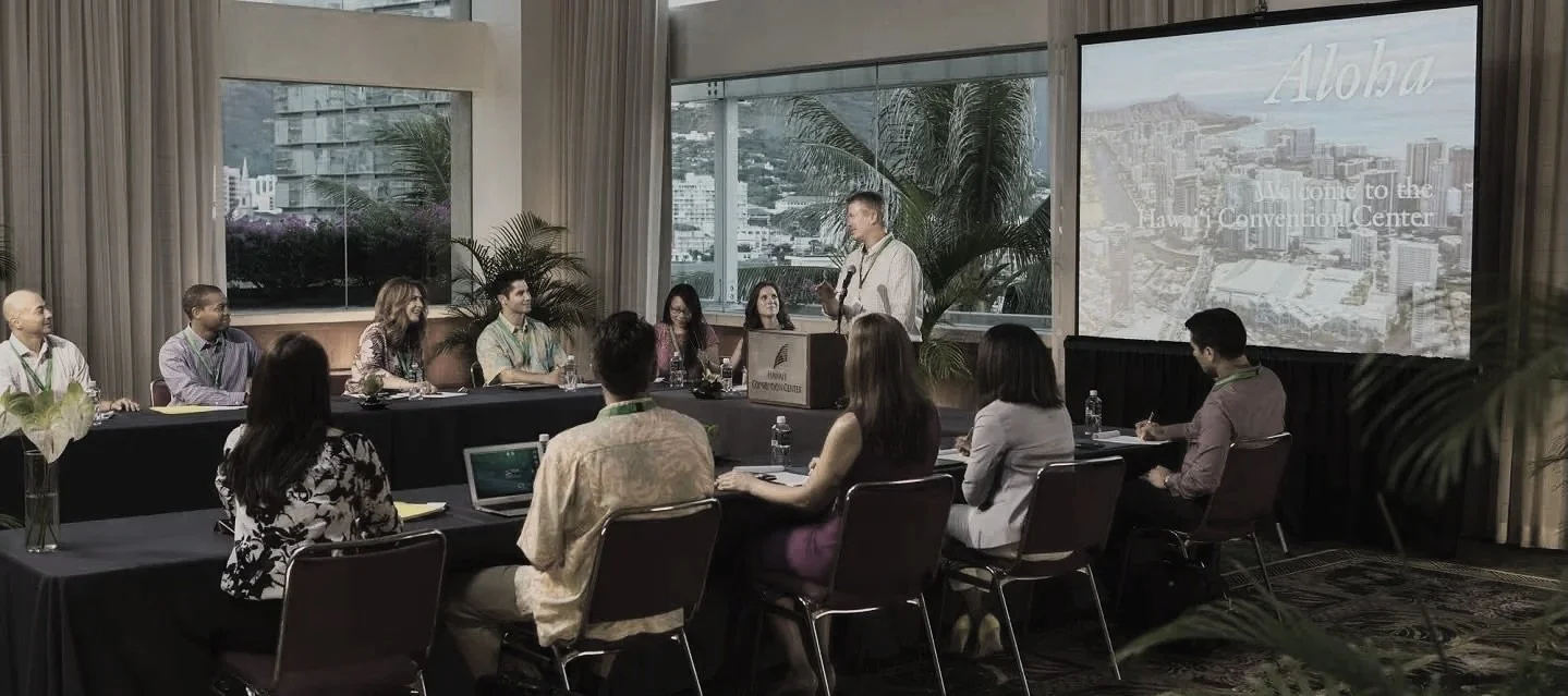 A man delivers a presentation at a conference table with seated attendees listening, large screen displaying 'Aloha' and a cityscape in the background.