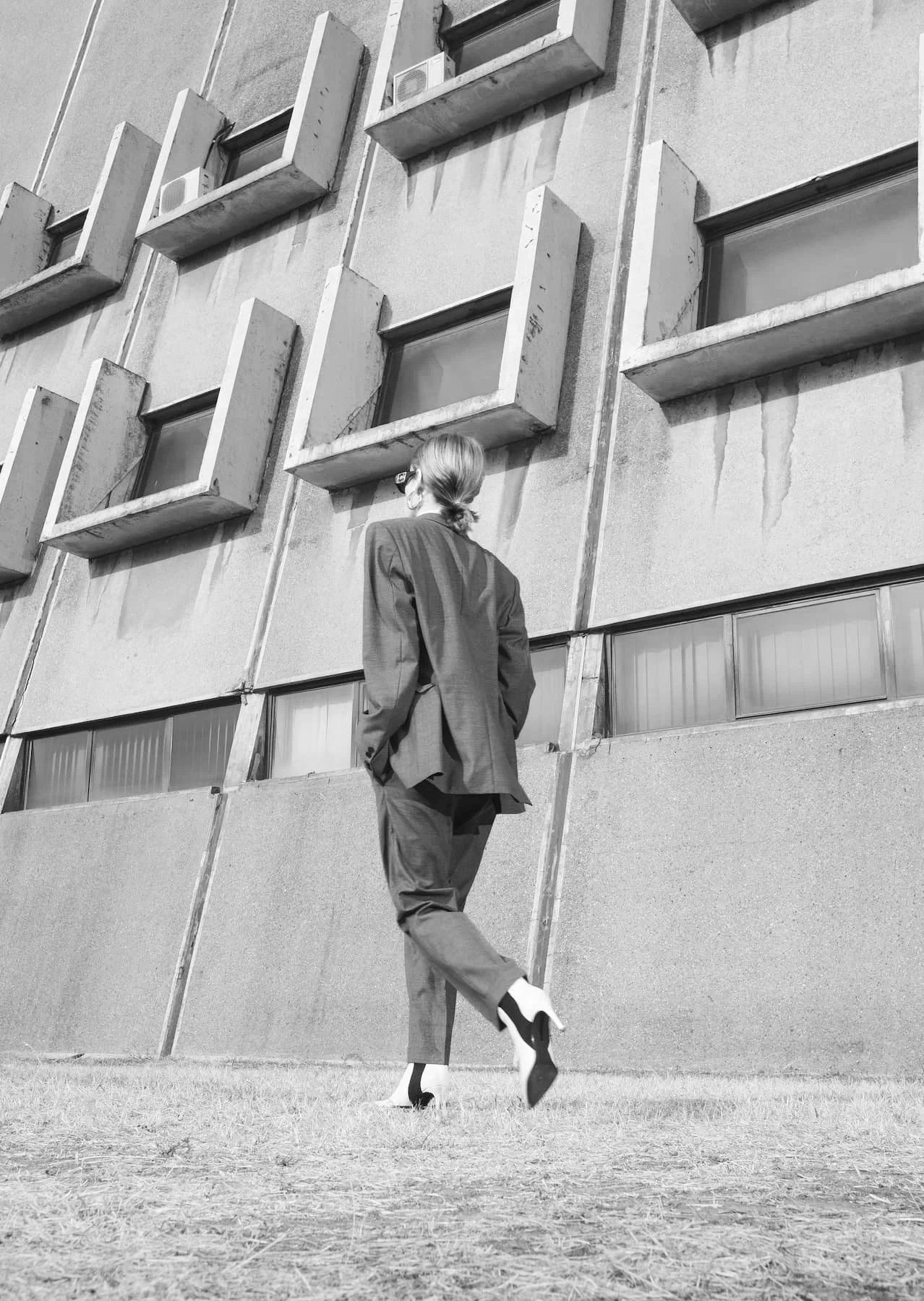 A woman in a business suit walking in front of a modern building with concrete walls and rectangular windows.