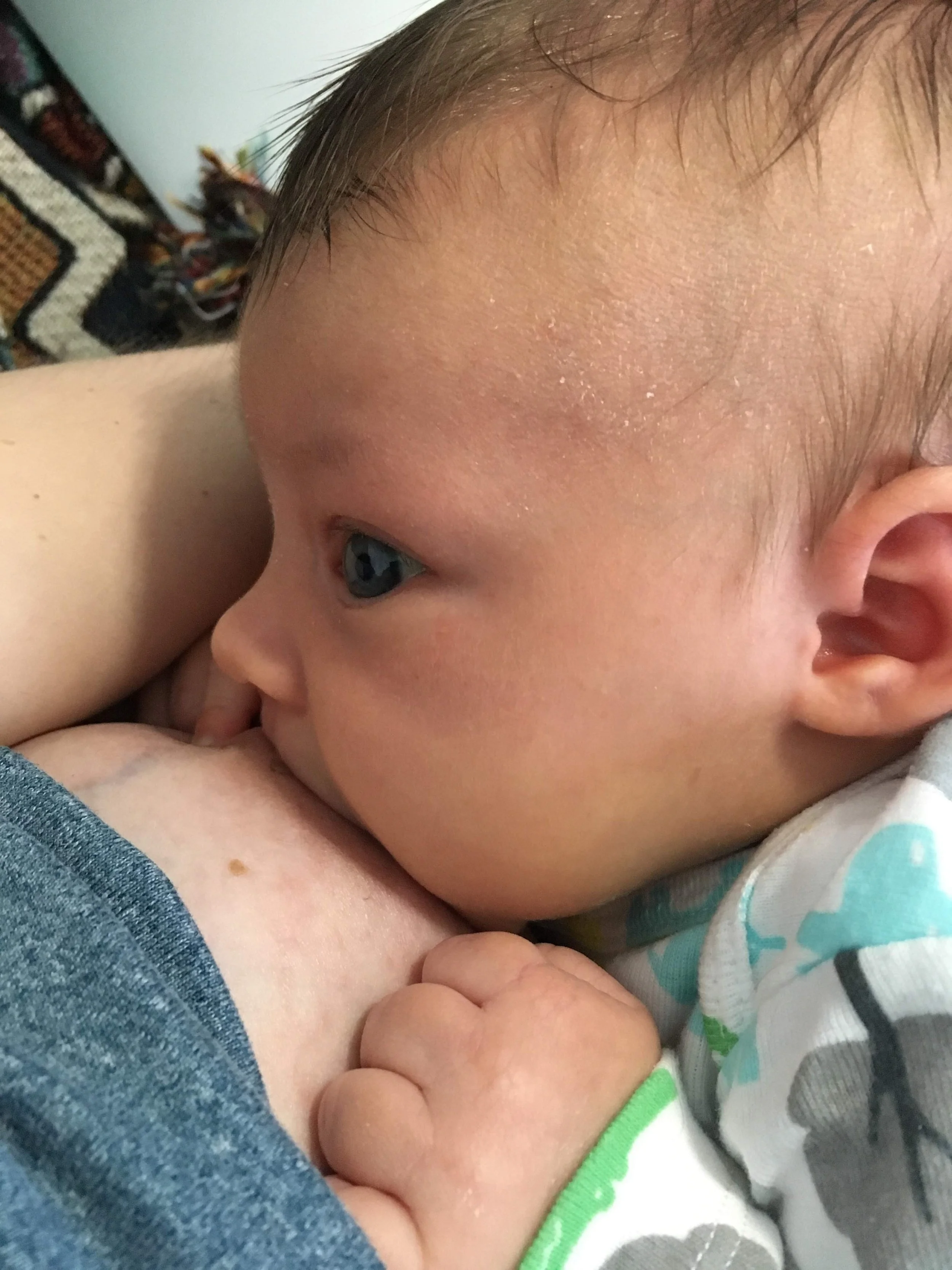 Close-up of a breastfeeding newborn, showing part of their forehead, eye, and ear, with a finger near their cheek, wearing a striped shirt with green accents.