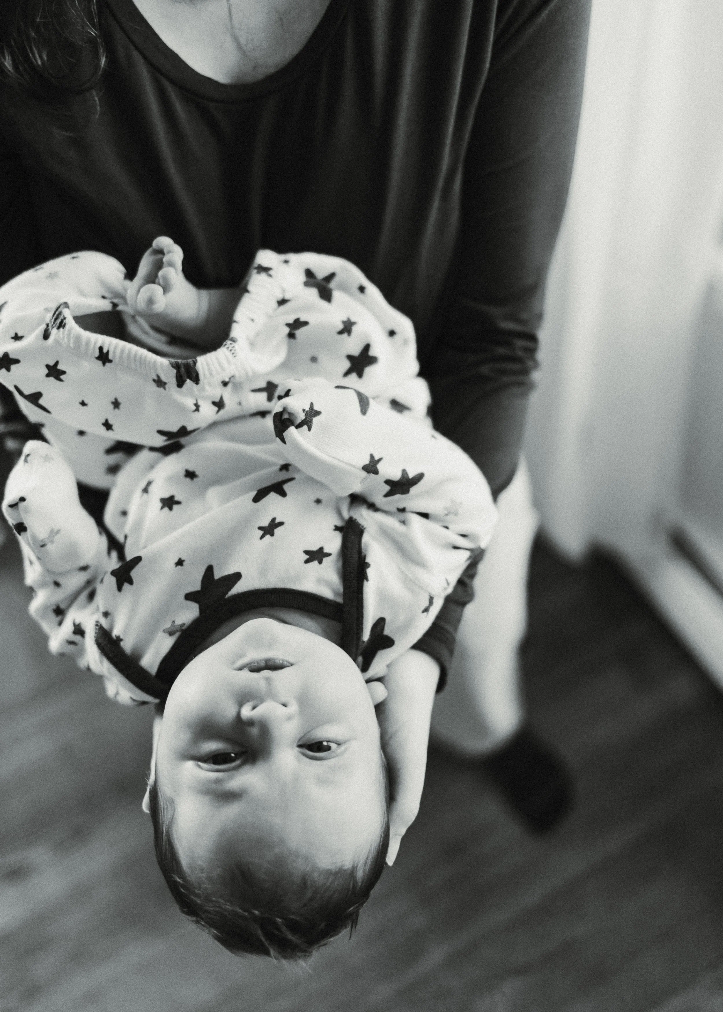 A young child with short hair is lying upside down on the floor, smiling, with his head closest to the camera. He is wearing a white shirt with star and bow patterns. An adult, dressed in a black shirt, is partially visible, supporting the child's back as he lies on his stomach on the floor.
