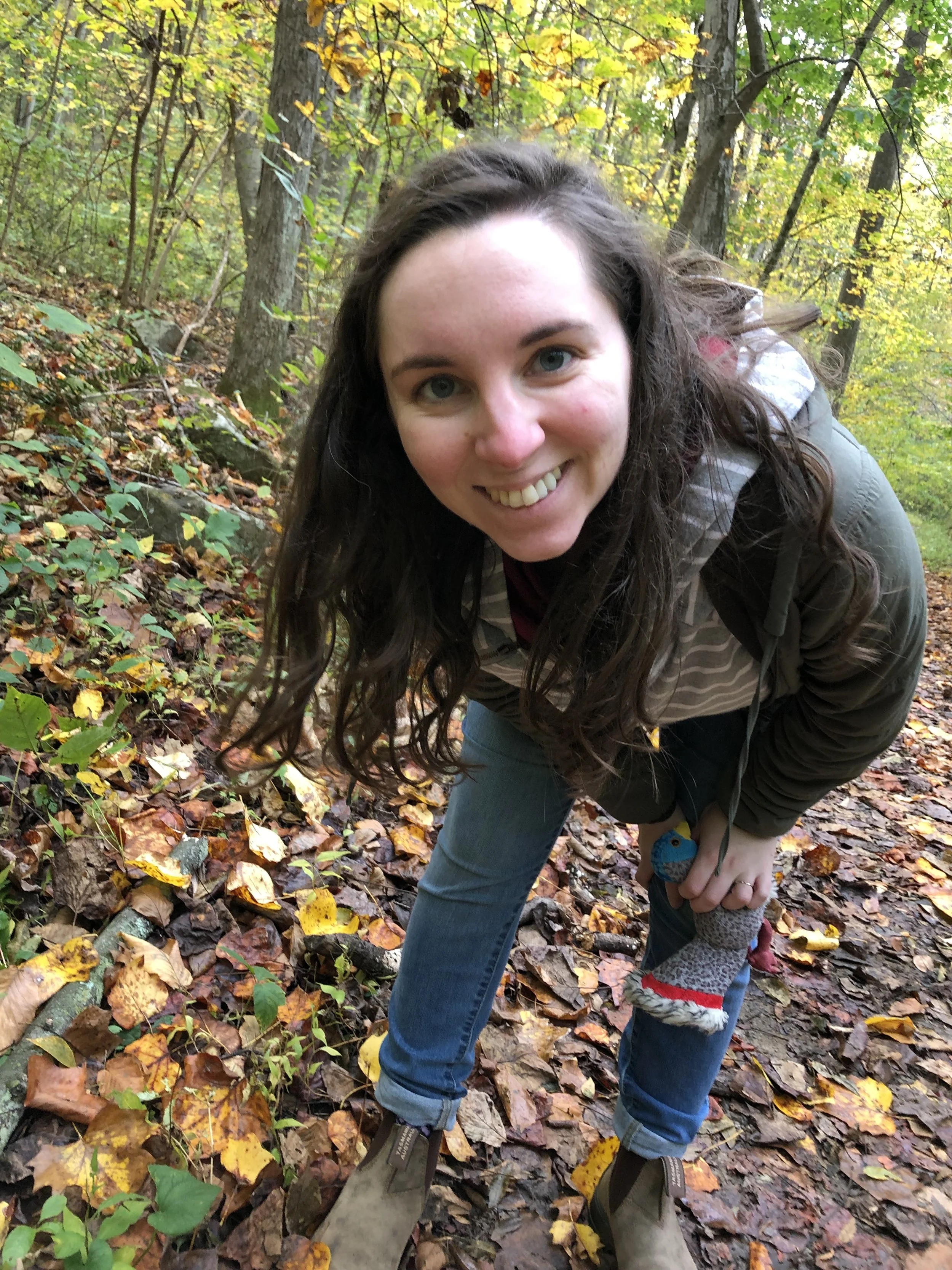 A woman with long curly hair smiling outdoors in a fall forest, wearing a jacket and jeans, standing on a leaf-covered trail.