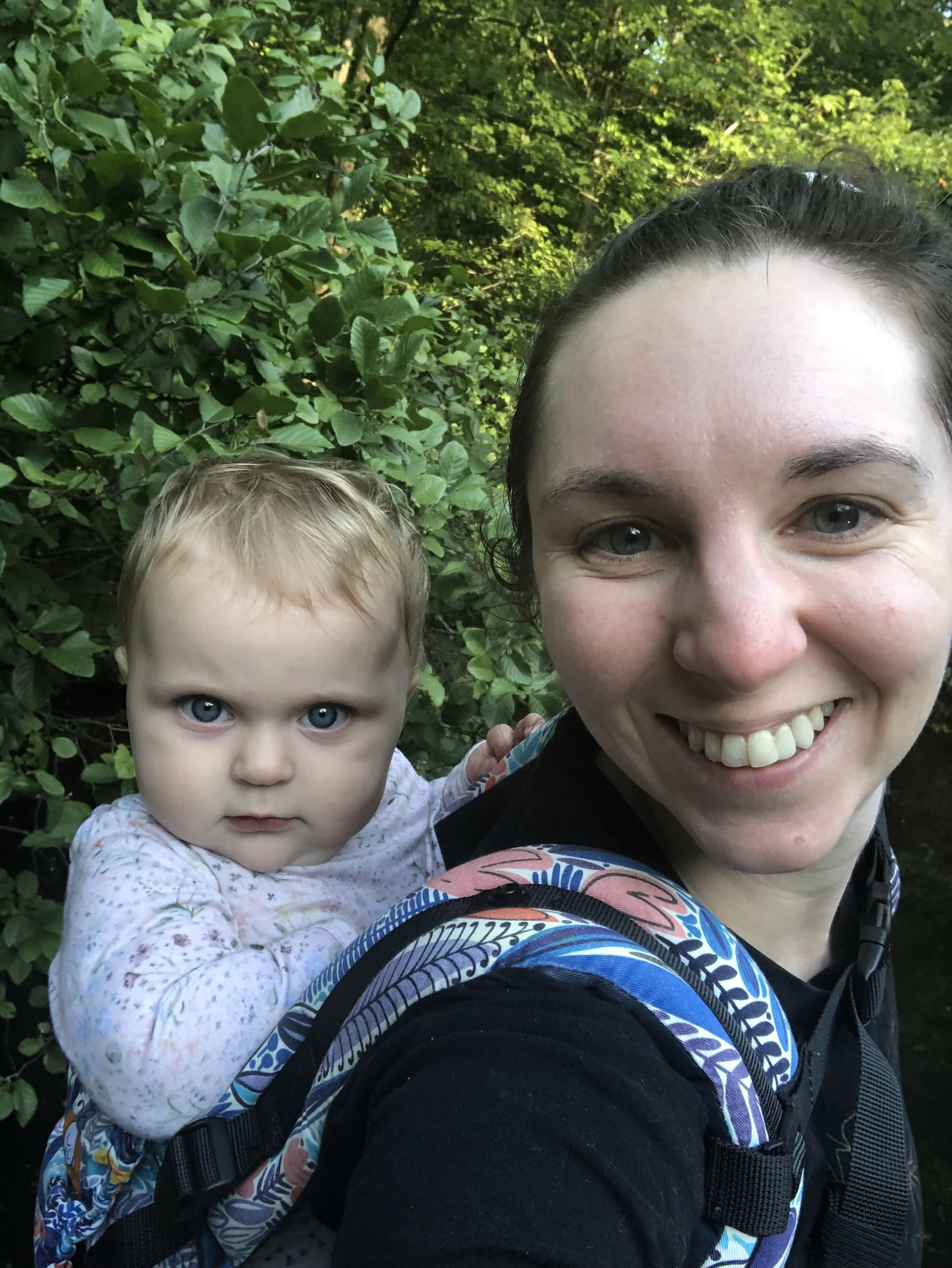 A woman smiling joyfully and a young girl in a backpack, both outdoors surrounded by green foliage.