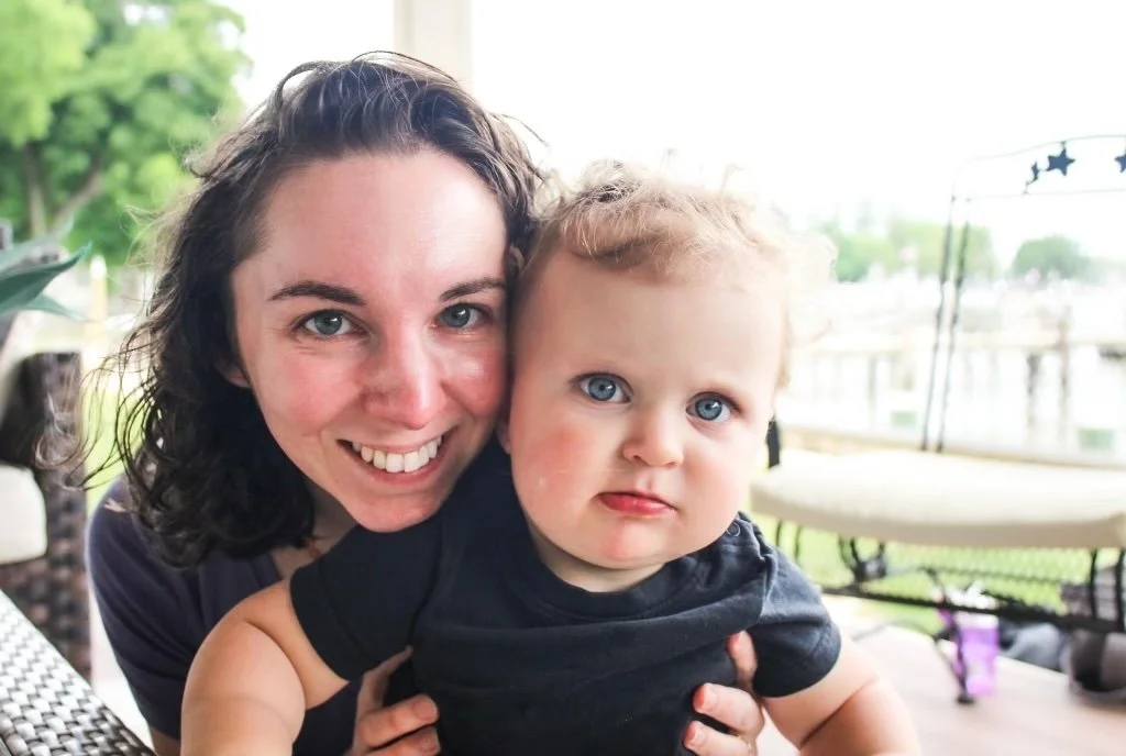 A woman with dark, curly hair smiling and holding a young child with curly blonde hair and blue eyes outdoors on a porch or patio.