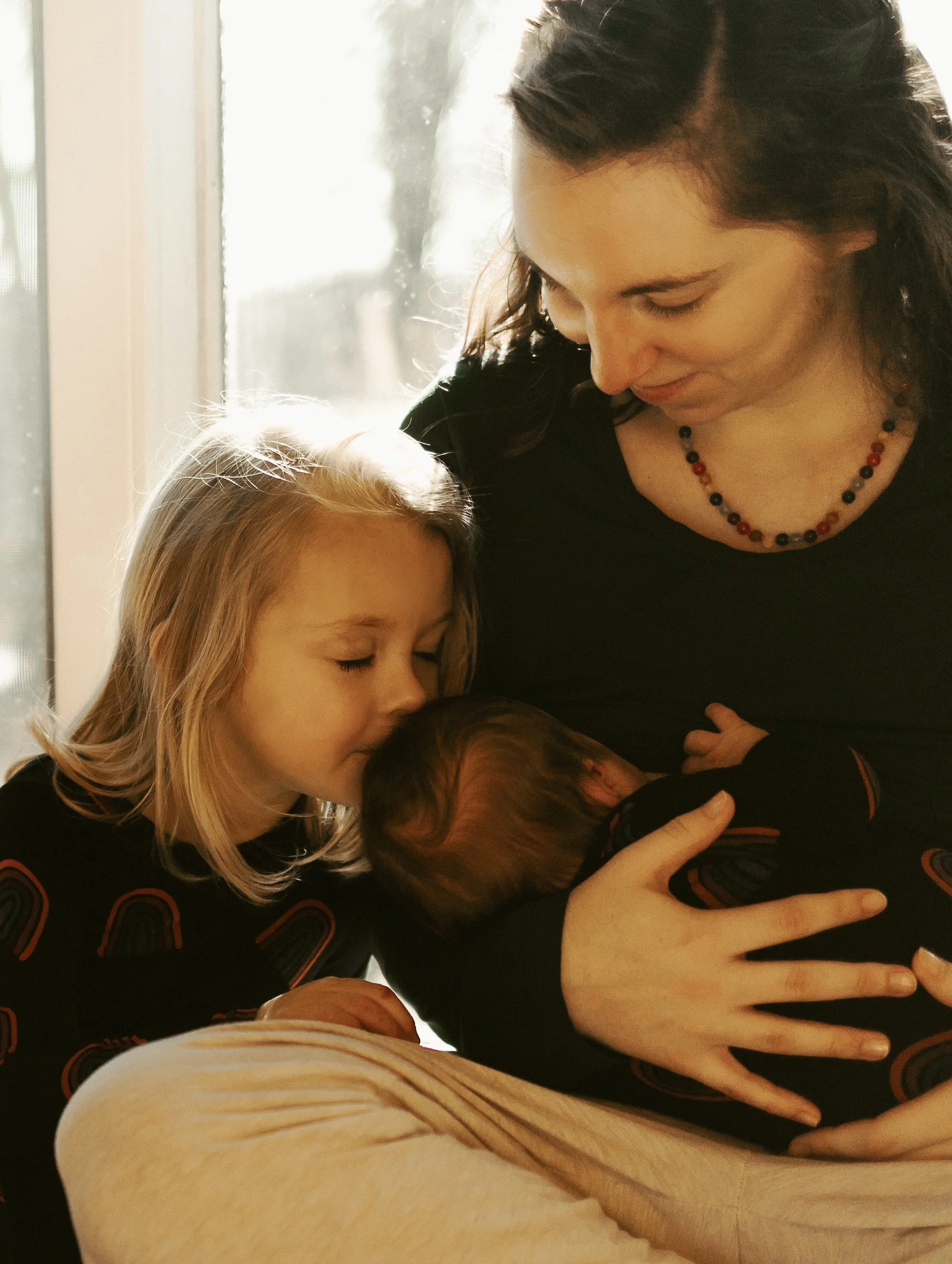 A woman with dark hair wearing a black shirt and colorful beaded necklace is holding and cuddling a baby while a young girl with blonde hair leans in close and kisses the baby on the head. They are near a window with sunlight shining through.