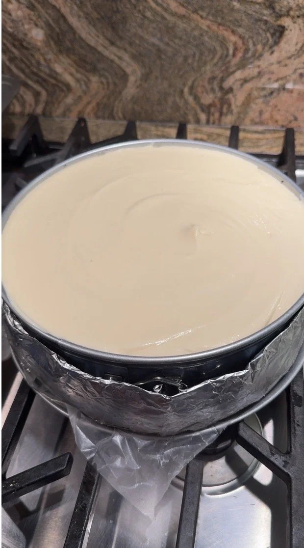“A close-up of smooth white‑chocolate cheesecake batter being spooned over the set chocolate layer inside a round baking pan.