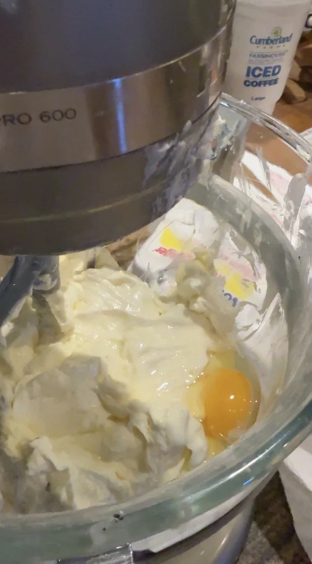 A close-up of a mixing bowl with softened cream cheese, sugar, and a cracked egg yolk being combined with a metal spatula.