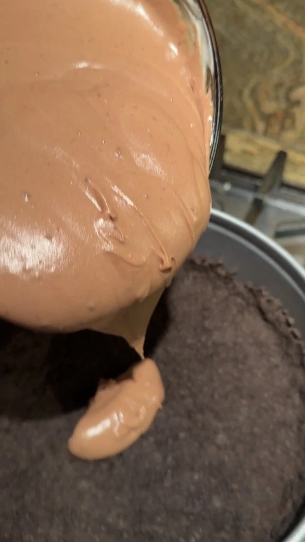 A close-up of thick chocolate cheesecake batter being spooned onto a pressed chocolate cookie crumb crust inside a round baking pan.