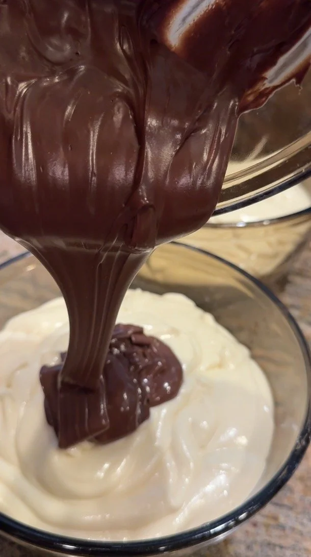 A close-up of melted milk chocolate streaming into a bowl of smooth cheesecake batter, creating a rich chocolate layer for the filling.