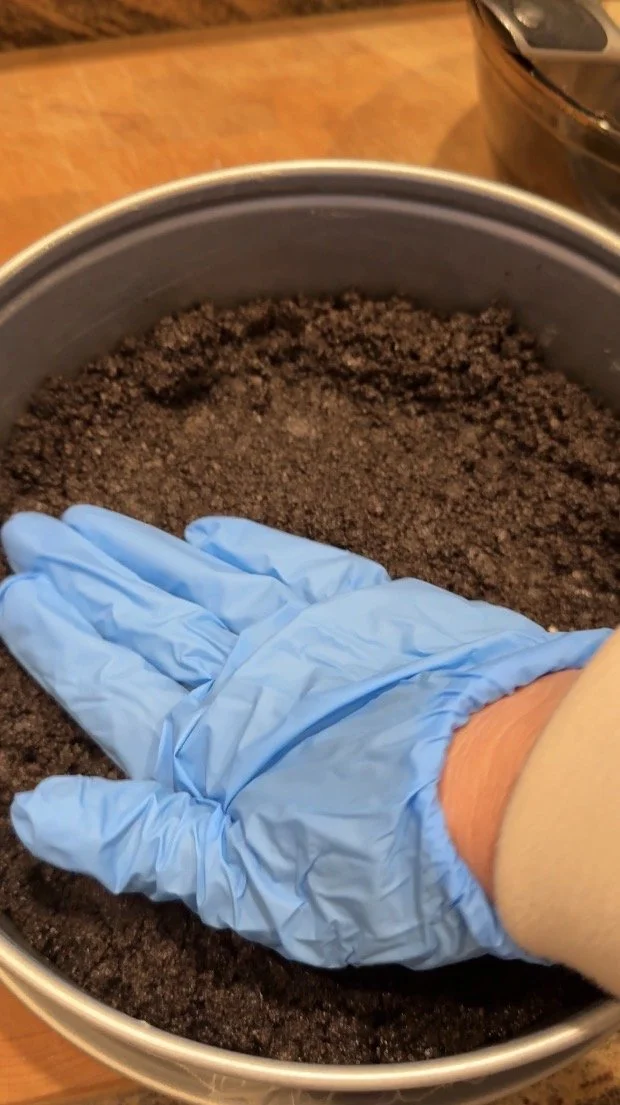 A close-up of a blue‑gloved hand pressing dark chocolate cookie crumbs into a pan to form a cheesecake crust.