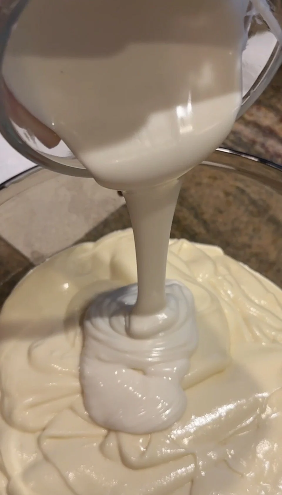 A close-up of melted white chocolate being poured into a bowl of smooth cheesecake batter to create the white‑chocolate layer.