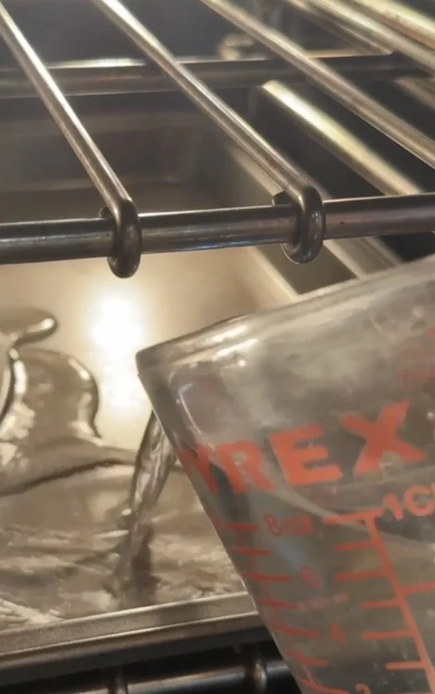 A close-up of water being poured from a glass measuring cup into a shallow pan that sits on the rack beneath the cheesecake to create gentle oven steam.