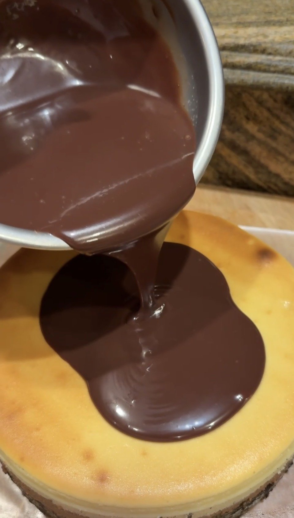 A close-up of glossy melted chocolate ganache being poured from a container onto the top of a fully chilled cheesecake.