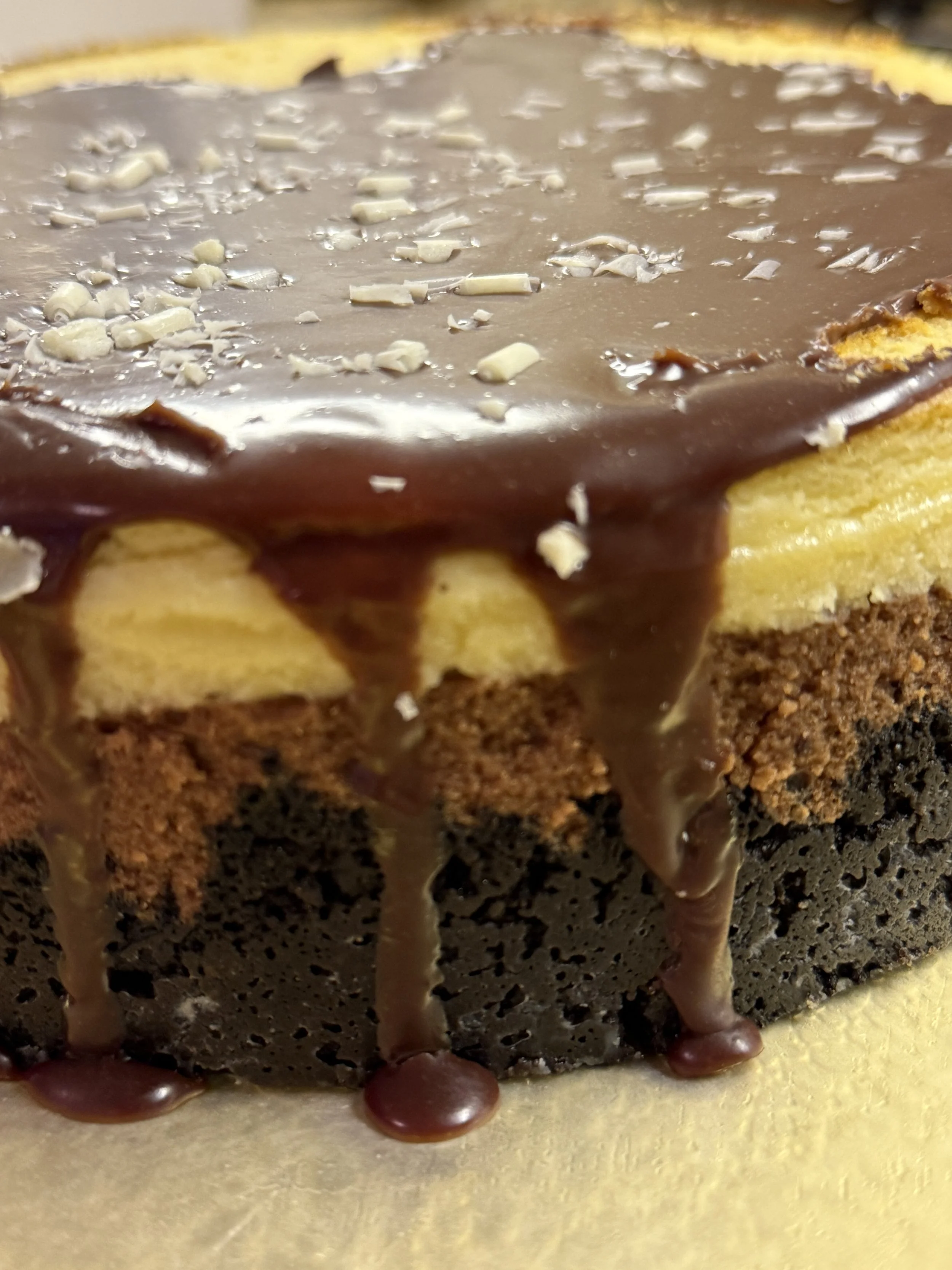 “A close-up of a finished triple‑layer cheesecake topped with smooth chocolate ganache that drips down the sides, with white‑chocolate shavings scattered across the top.