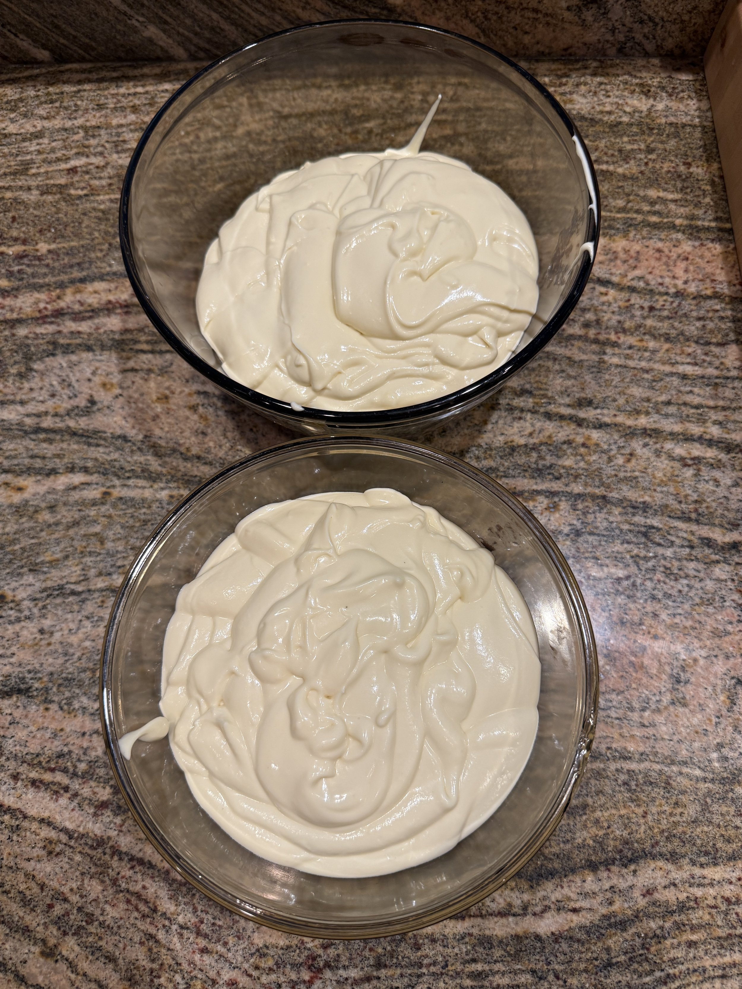 Two glass bowls sitting on a countertop, each holding a portion of smooth, creamy cheesecake batter after being divided for the chocolate layers.