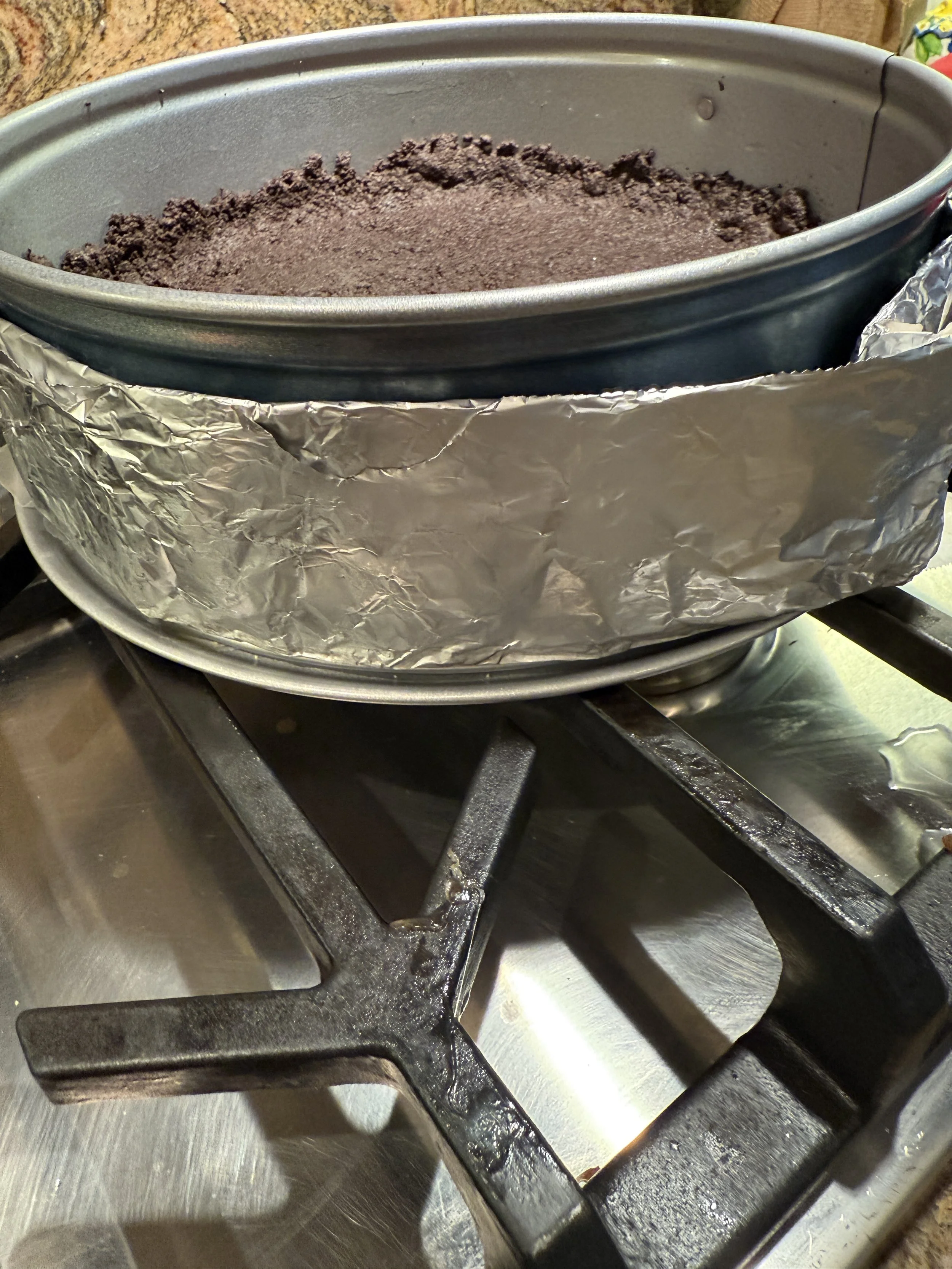 A springform pan wrapped on the outside with a damp paper‑towel and aluminum foil collar, holding a pressed chocolate cookie crumb crust.