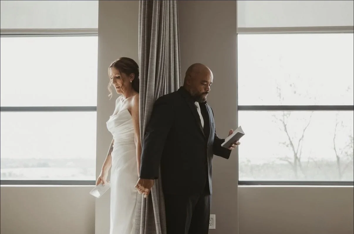A bride in a white wedding dress and a groom in a black tuxedo hold hands behind a curtain in front of large windows, with the bride holding a glass and the groom reading a small book.