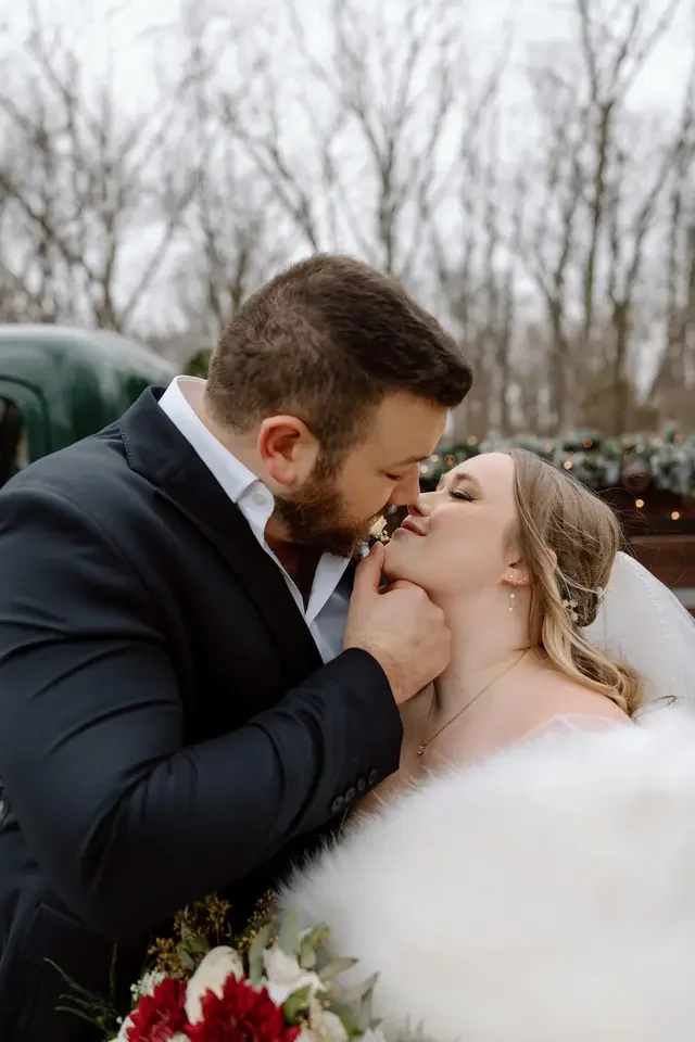 A bride and groom sharing a romantic moment outdoors, with trees in the background, the groom gently holding the bride's chin as they lean in for a kiss, the bride wearing a white fur cover and holding a bouquet.