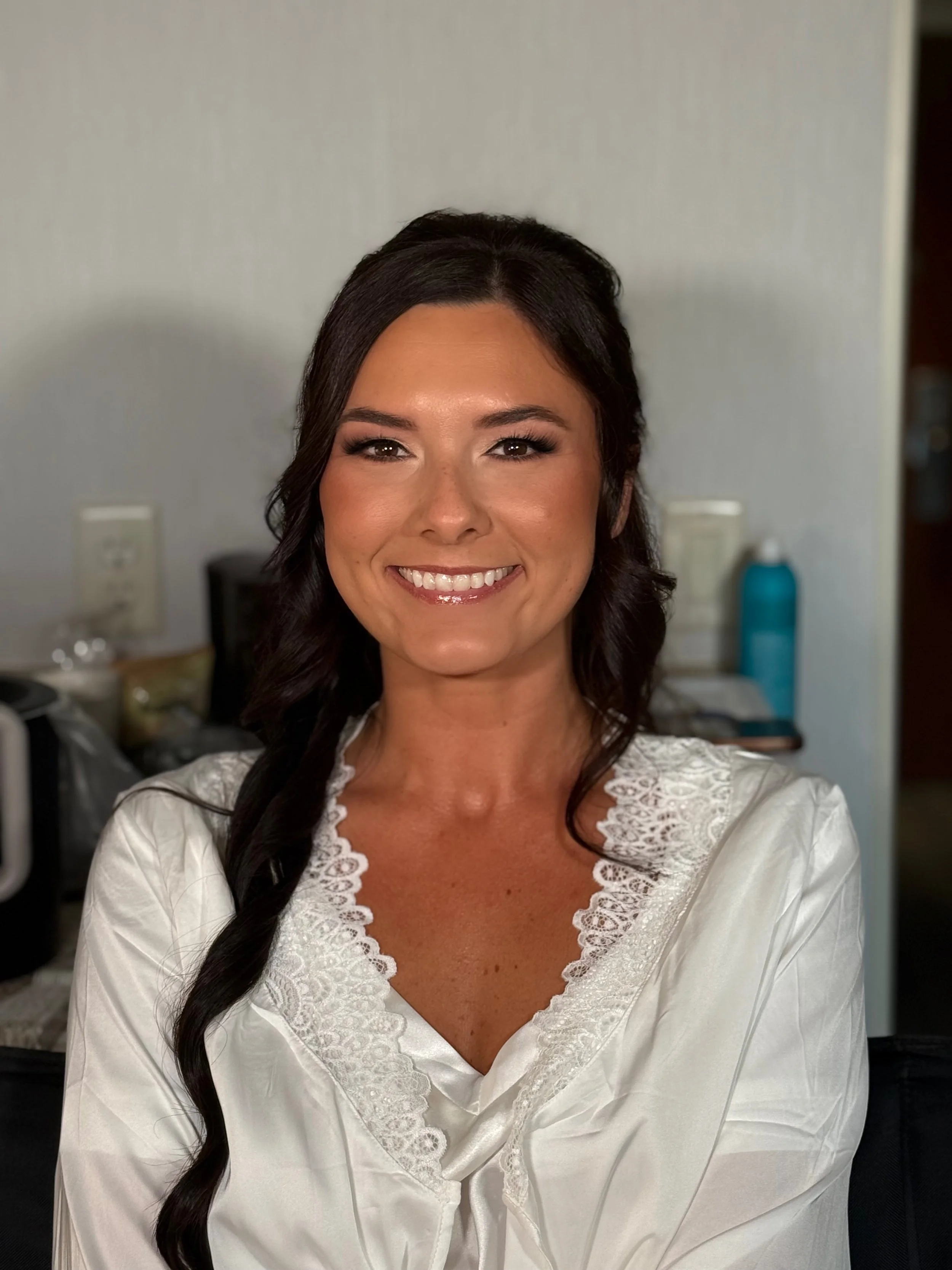 A woman with long dark hair styled in loose curls, smiling in a white satin top with lace details.