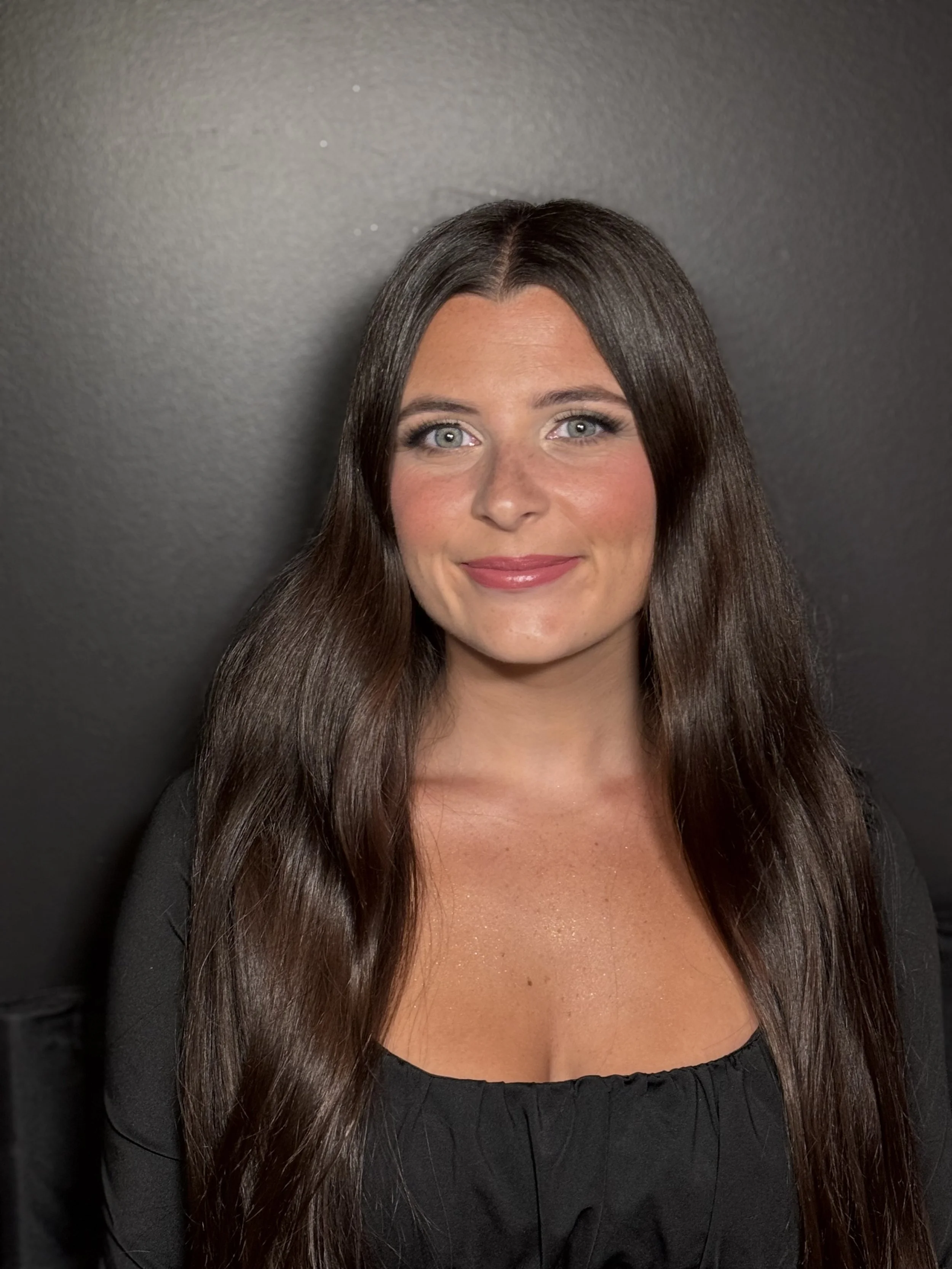 Portrait of a woman with long brown hair, blue eyes, and a black top, smiling against a dark gray background.