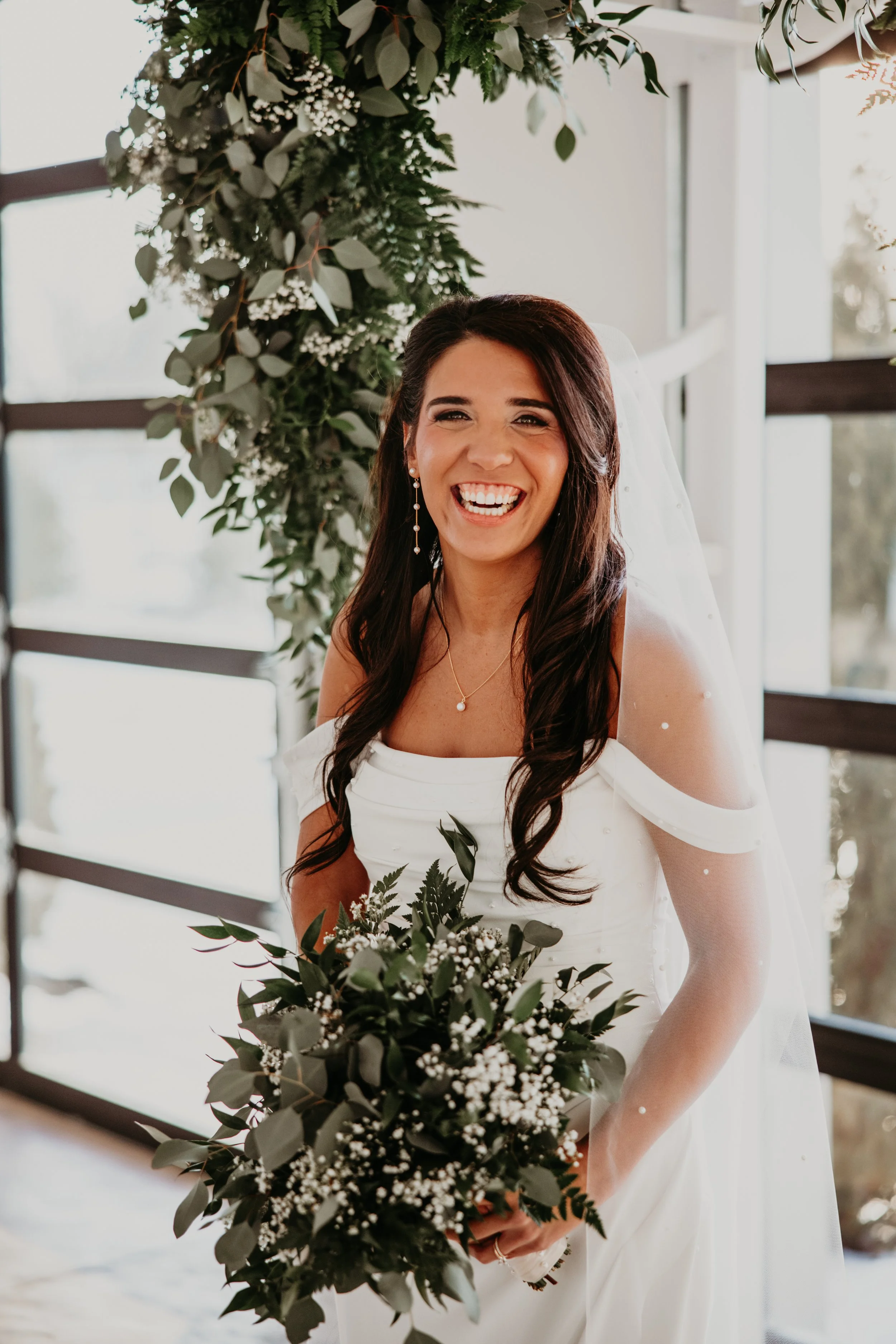 A joyful bride with dark hair in a white wedding dress holding a bouquet of greenery and small white flowers, standing indoors with large windows and green foliage in the background.
