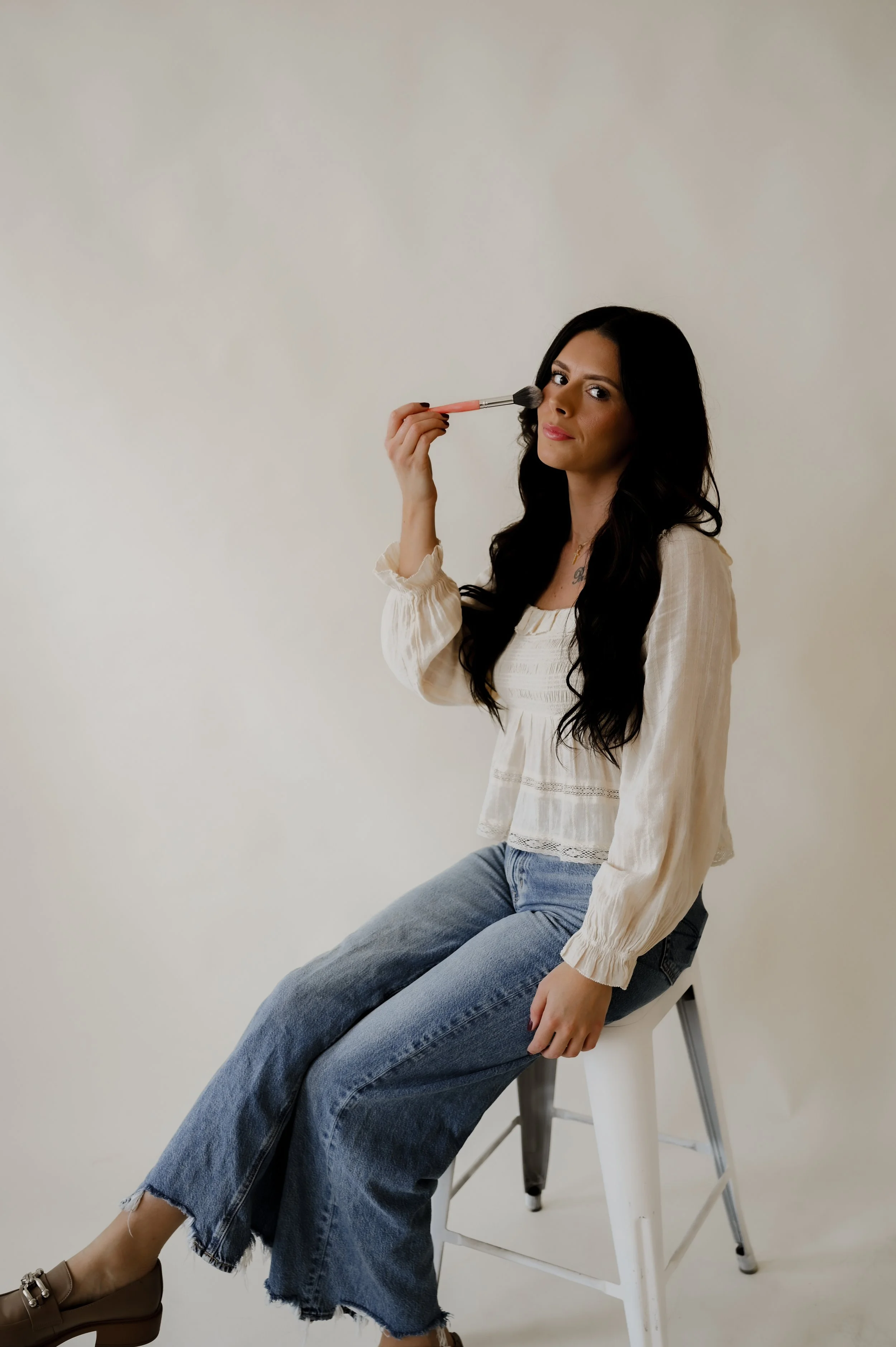 A woman with long dark hair sitting on a white stool, applying makeup with a brush, wearing a cream-colored blouse and blue jeans, against a plain light background.