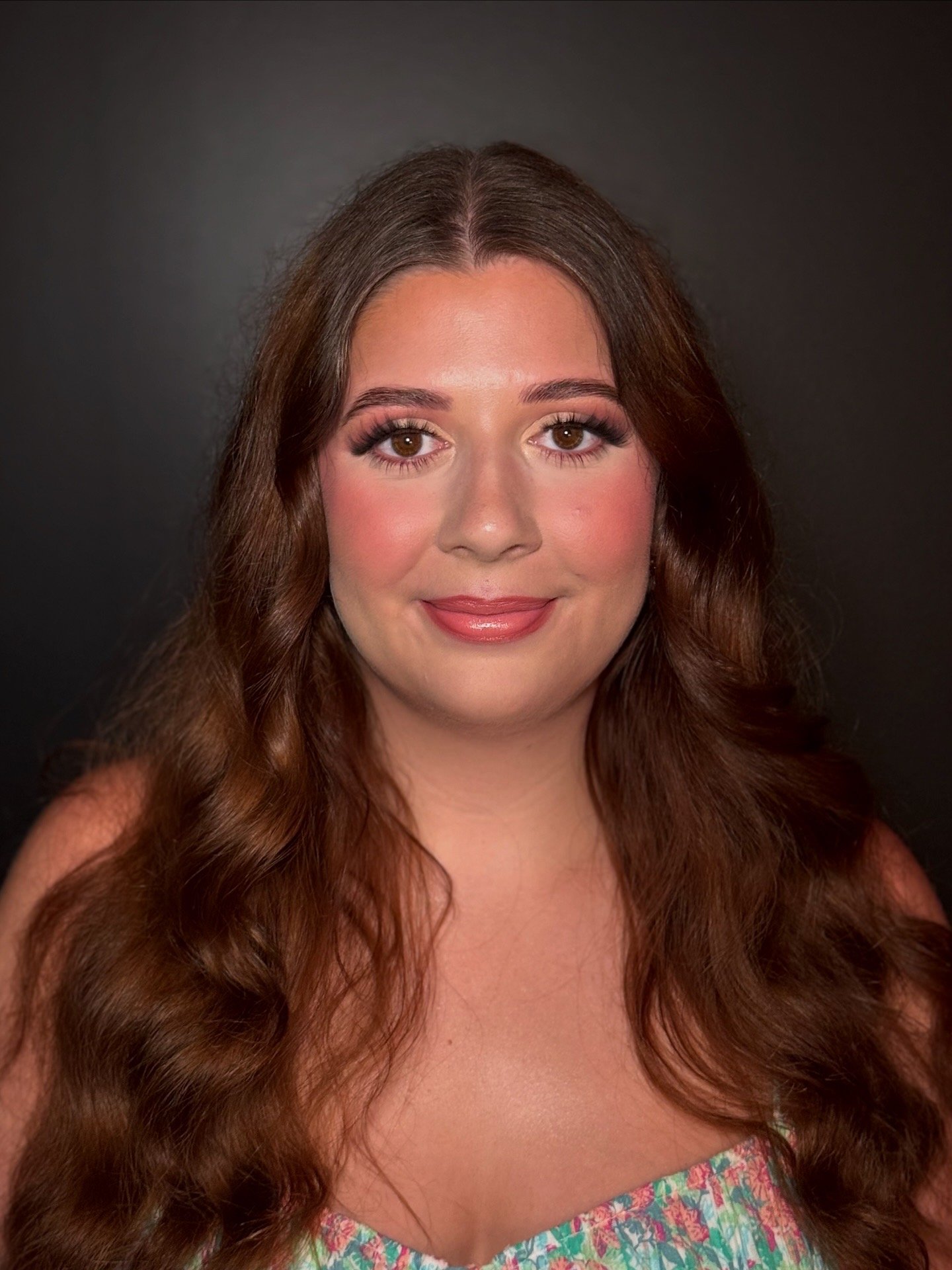 Portrait of a woman with long wavy brown hair, wearing makeup, a warm smile, and a floral dress against a dark background.