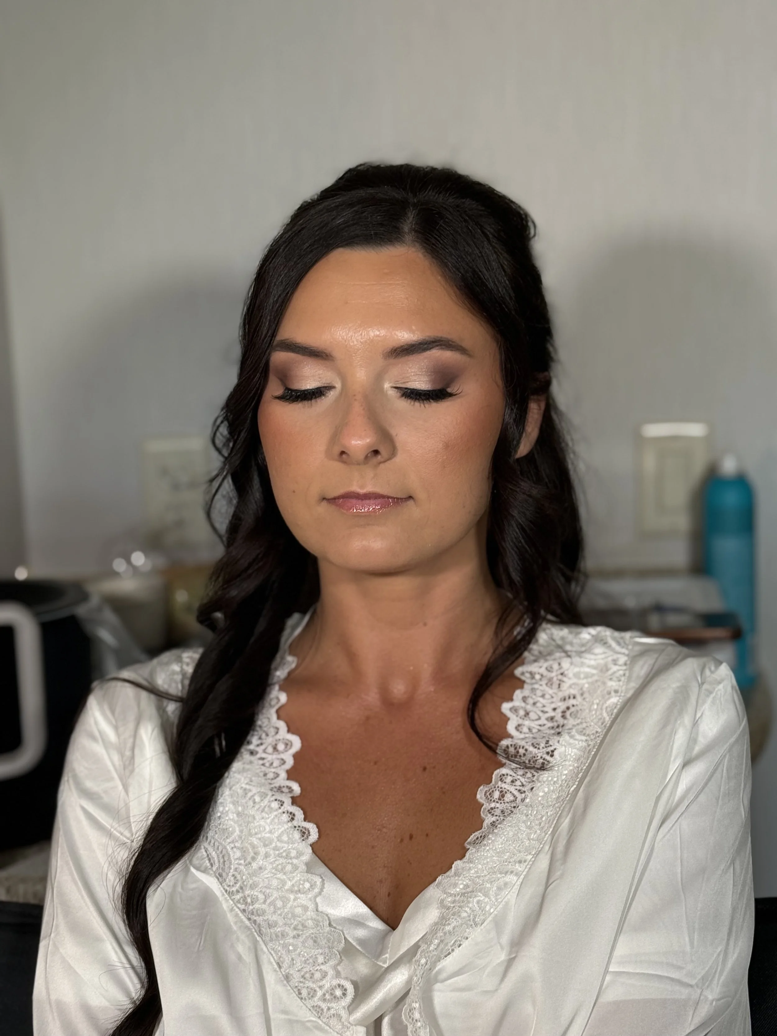 A woman with dark brown hair styled in loose curls, wearing a white garment with lace details, has her eyes closed and appears to be preparing for a makeup or skincare routine in a room with neutral-colored walls.