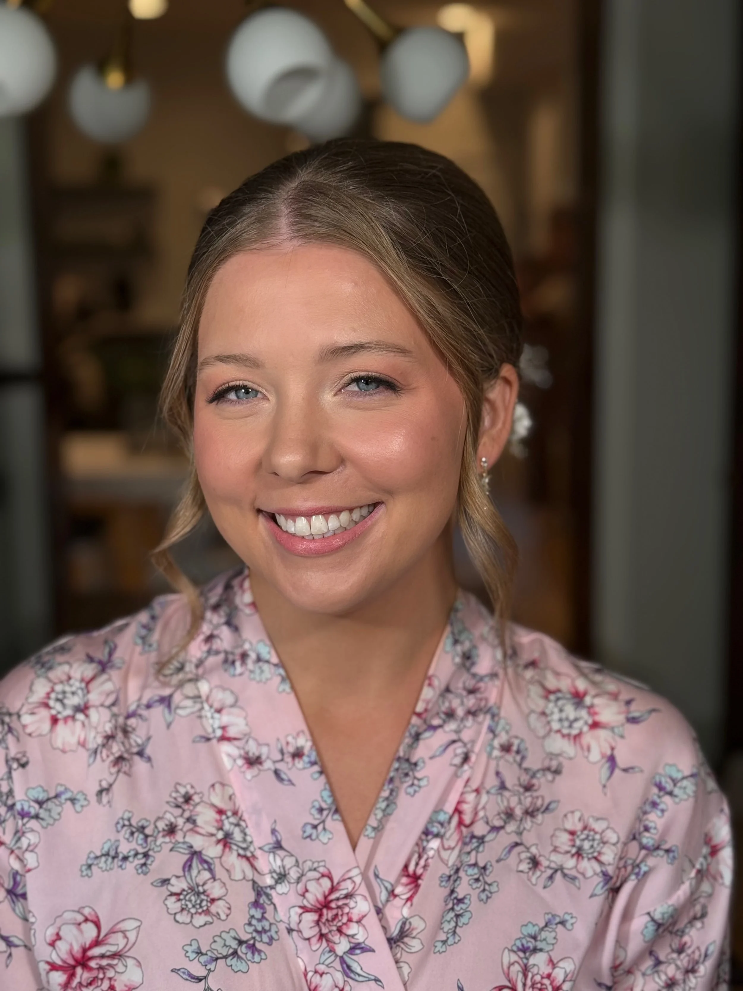 Close-up of a smiling woman with light brown hair styled in loose waves, wearing a pink floral kimono-style top, in a warmly lit indoor setting.