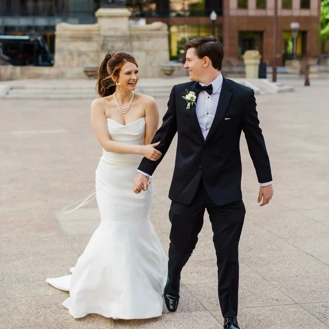 A newlywed couple in wedding attire walking outdoors and smiling, with the bride in a white strapless gown and the groom in a black tuxedo with a boutonniere.