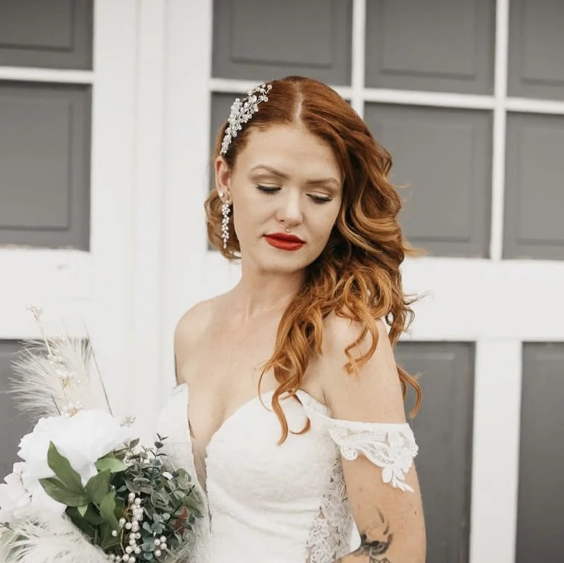 A woman with red hair and fair skin in a white wedding dress holding a bouquet of white flowers and greenery, standing outdoors in front of a gray barn door, with her eyes closed and a serene expression.