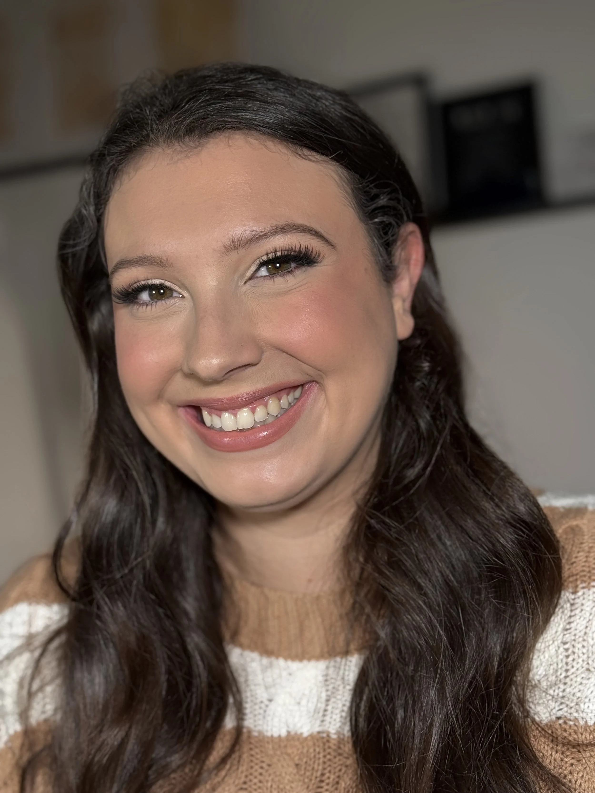 A woman with long dark brown wavy hair, light makeup, and a warm smile, wearing a beige and white striped sweater.