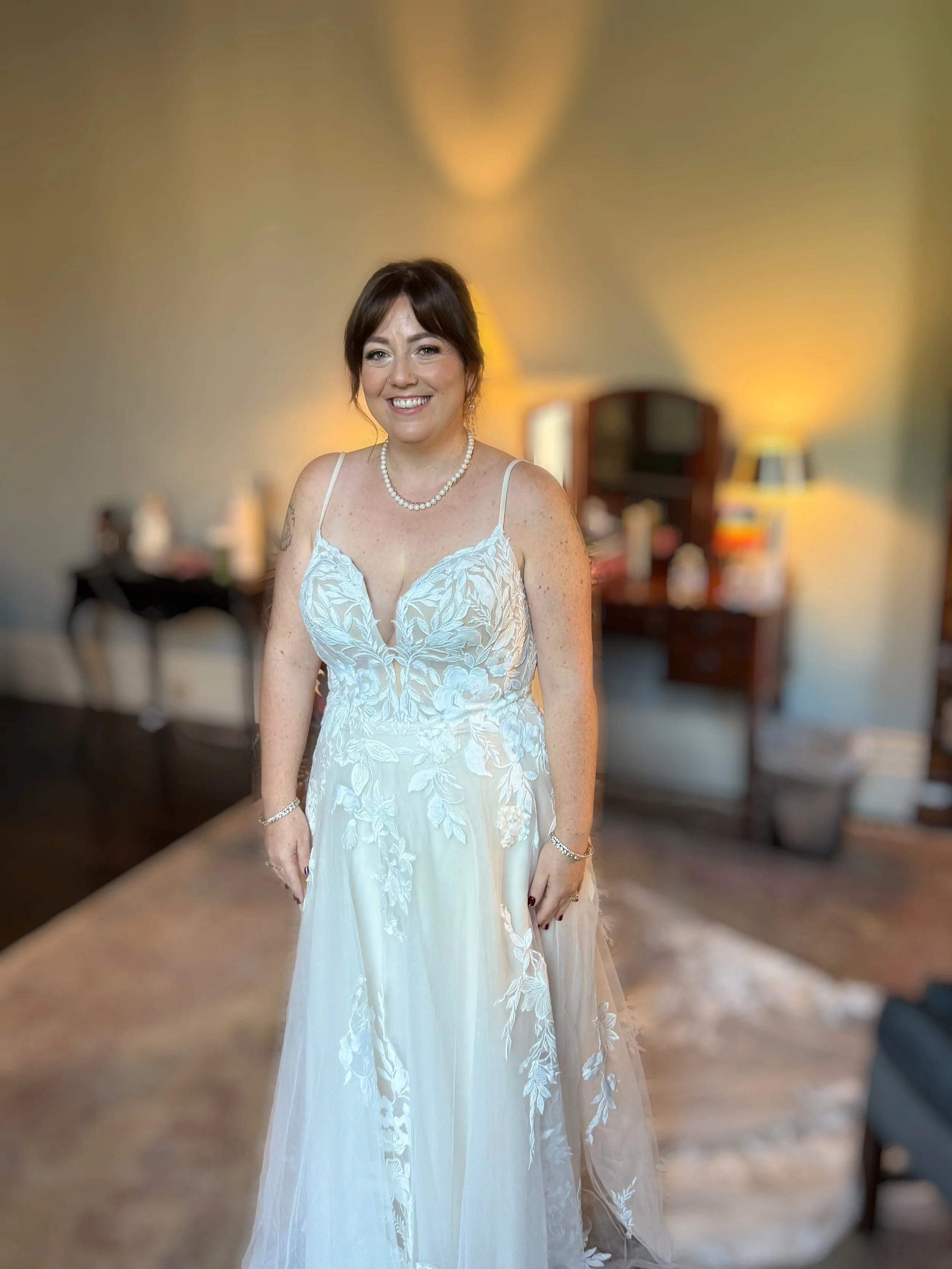 A woman in a white wedding dress with floral embroidery, wearing a pearl necklace, smiling in a warmly lit room.