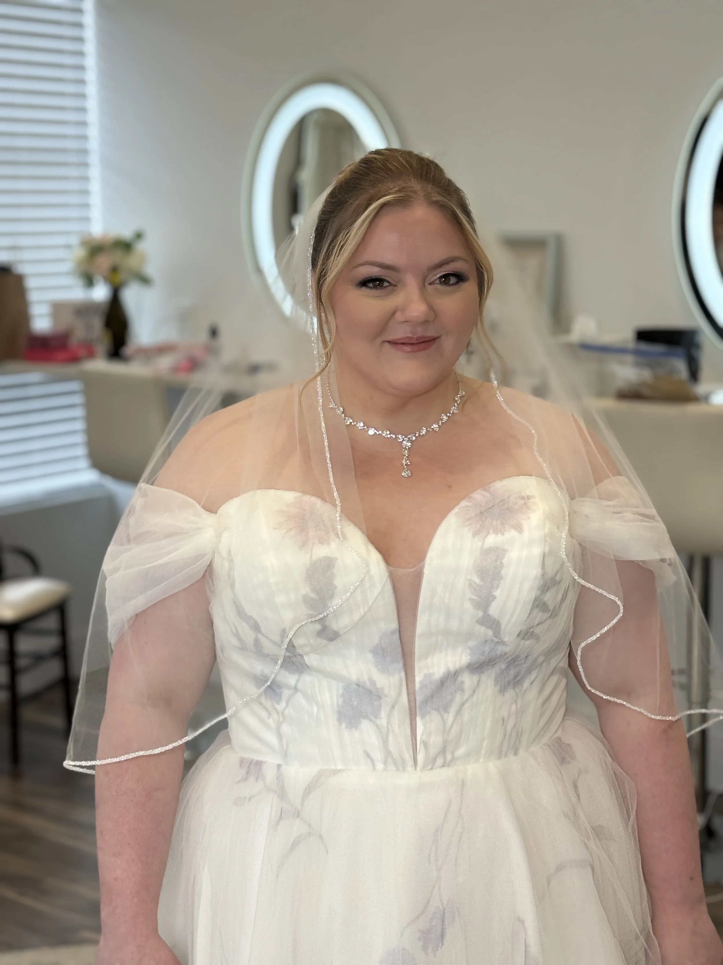 A bride wearing a white wedding dress with floral patterns, pearl necklace, and veil, smiling in a bridal room with mirrors and makeup items.