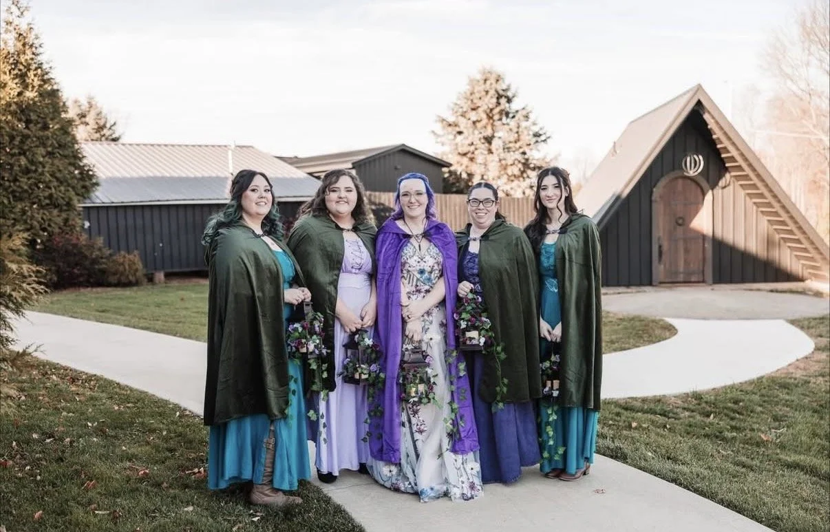 Five women dressed in fantasy-themed costumes standing outside on a sidewalk near a small black building with a triangular roof, surrounded by trees and grass.