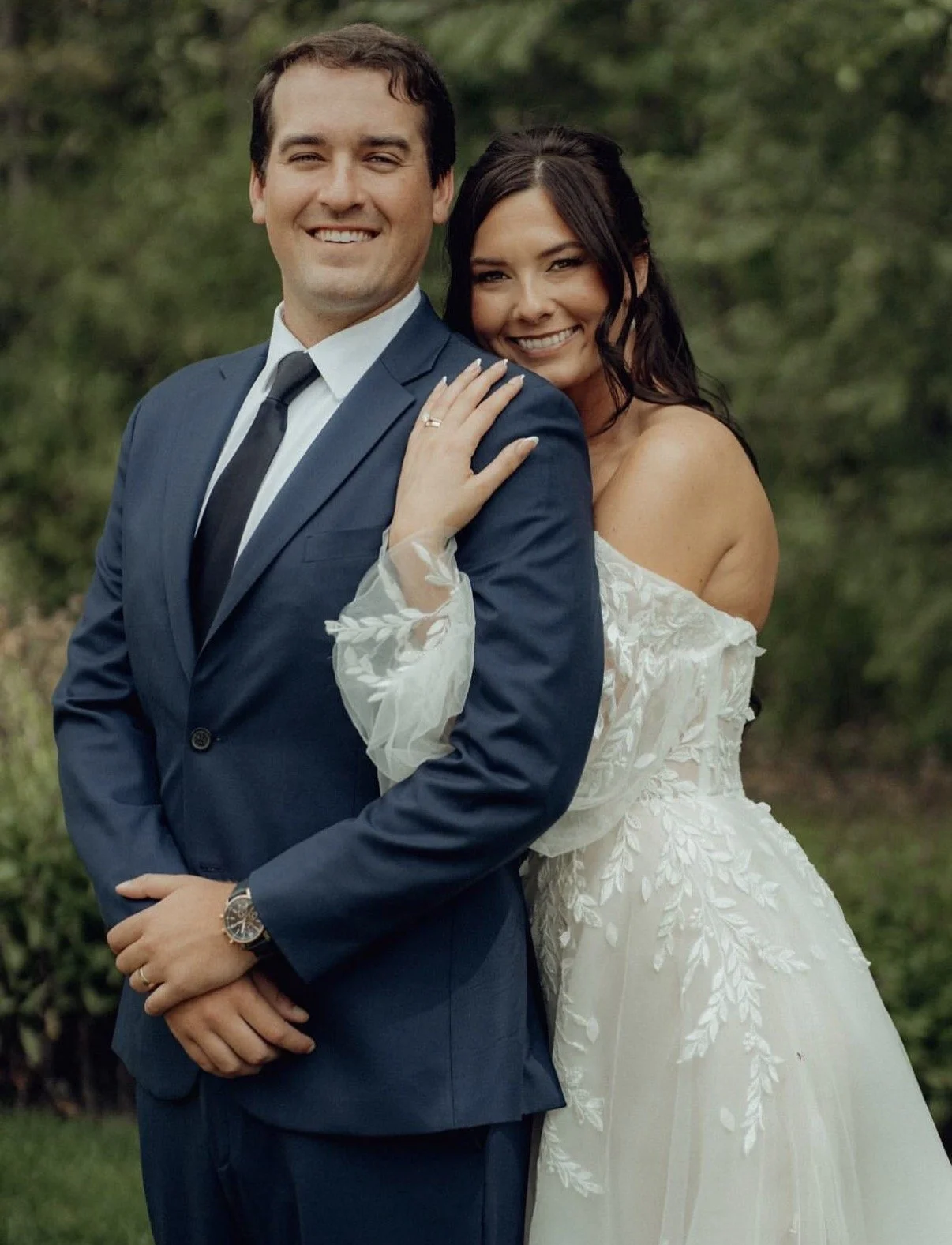 A smiling couple dressed in wedding attire outdoors, with the woman wearing a white off-shoulder wedding gown and the man in a dark blue suit, both posing happily in front of greenery.