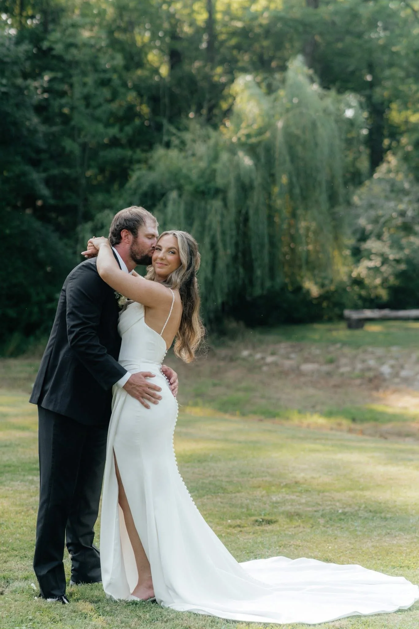 A bride and groom standing outdoors on a grassy area, embracing each other with big smiles, surrounded by green trees and foliage.