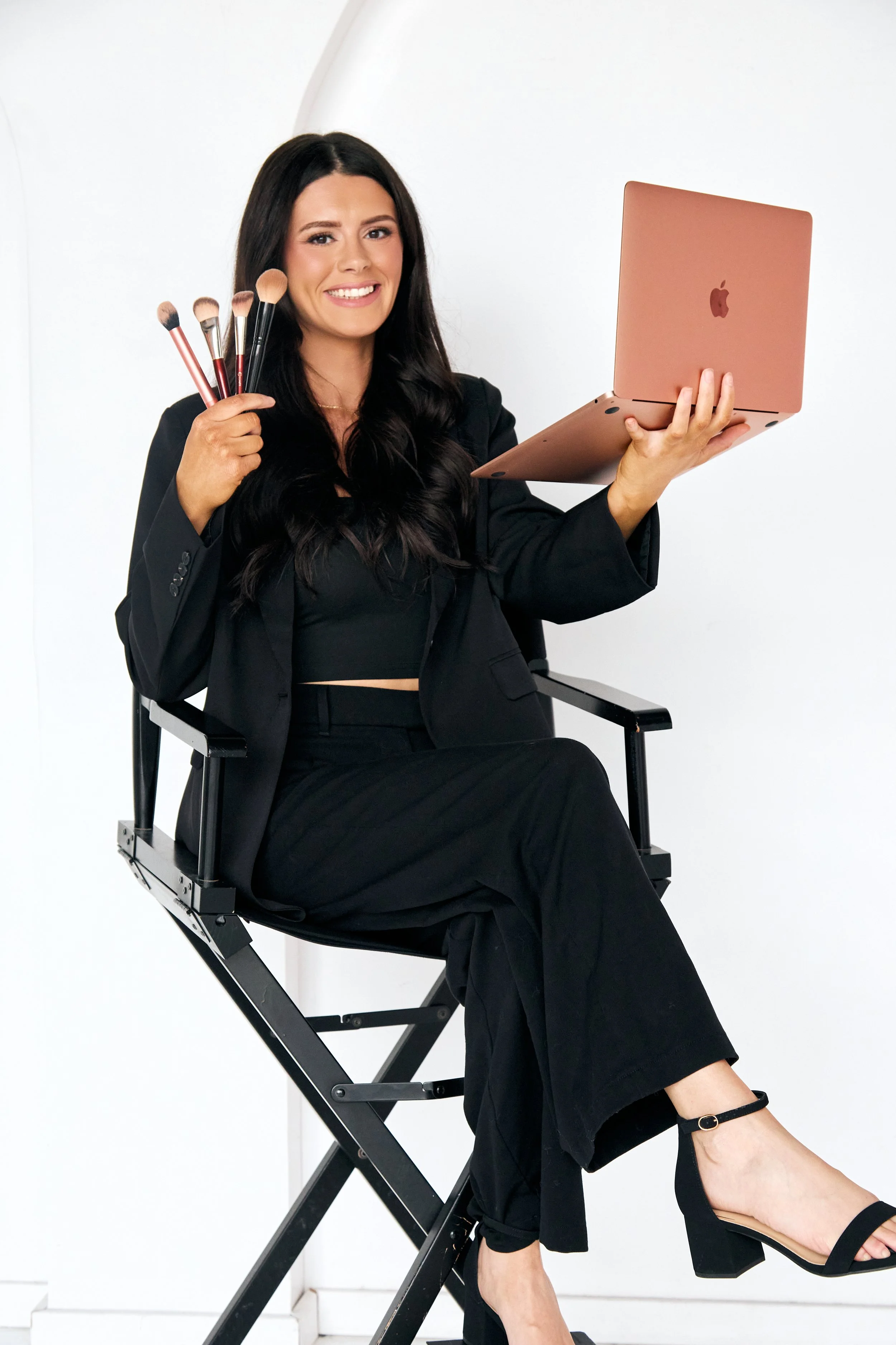 A woman sitting on a director's chair, holding a pink laptop in one hand and makeup brushes in the other, smiling at the camera. She is dressed in black with black heels, against a white background.