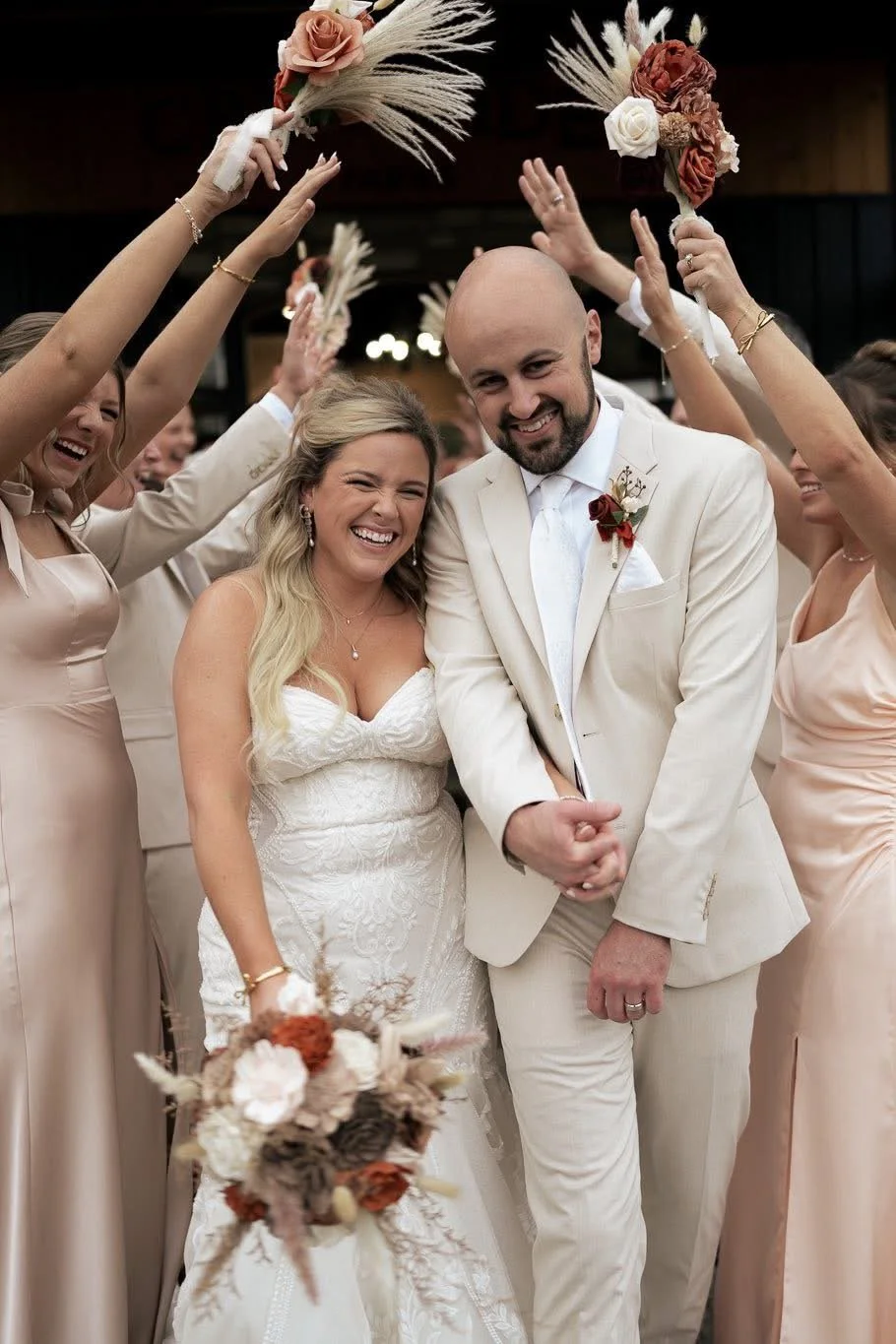 A bride and groom smiling at their wedding with bridesmaids holding bouquets over their heads, all dressed in light-colored attire in a joyful celebration.