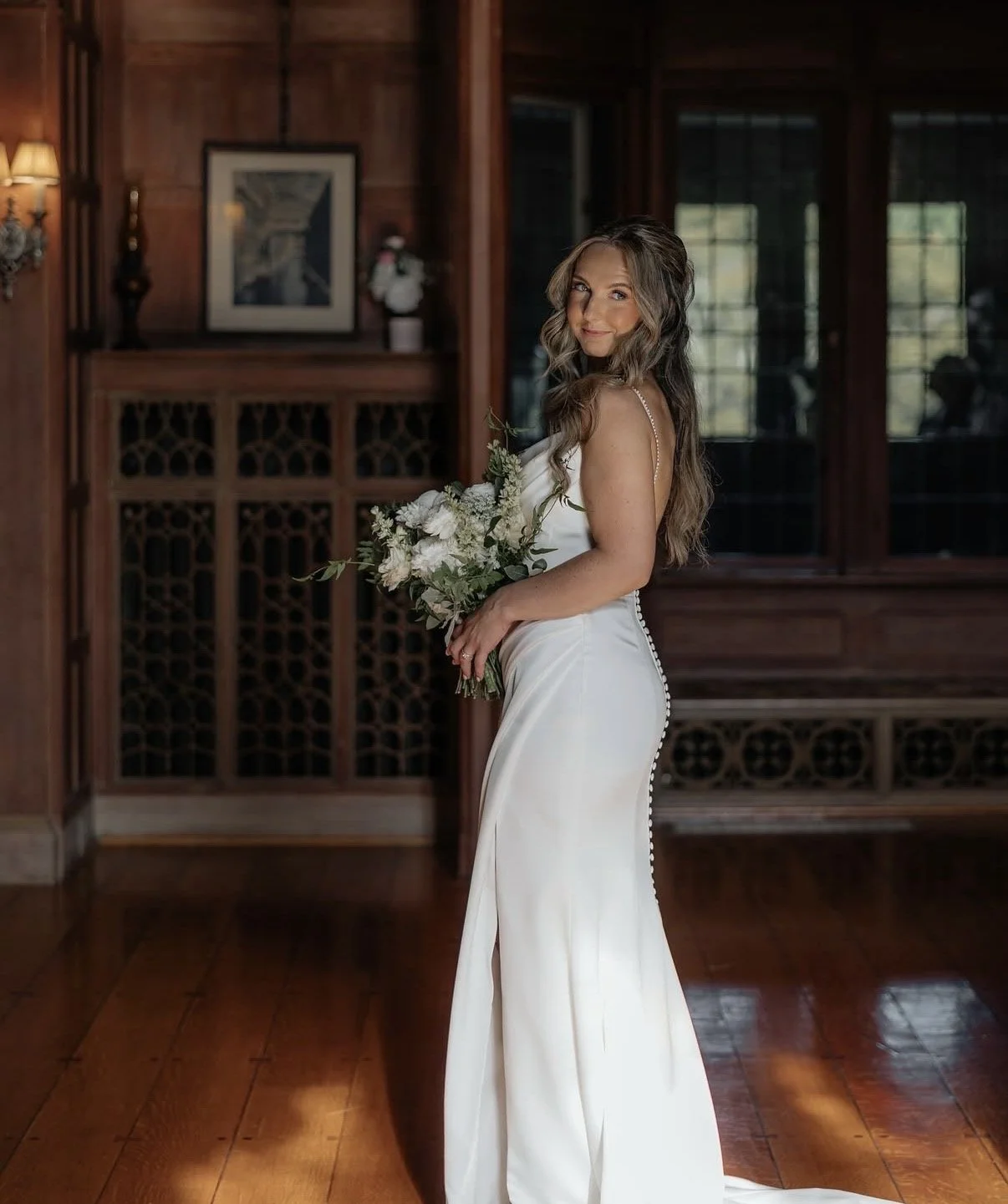 A bride in a white wedding gown holding a bouquet of white flowers, standing in a wood-paneled room with large windows.