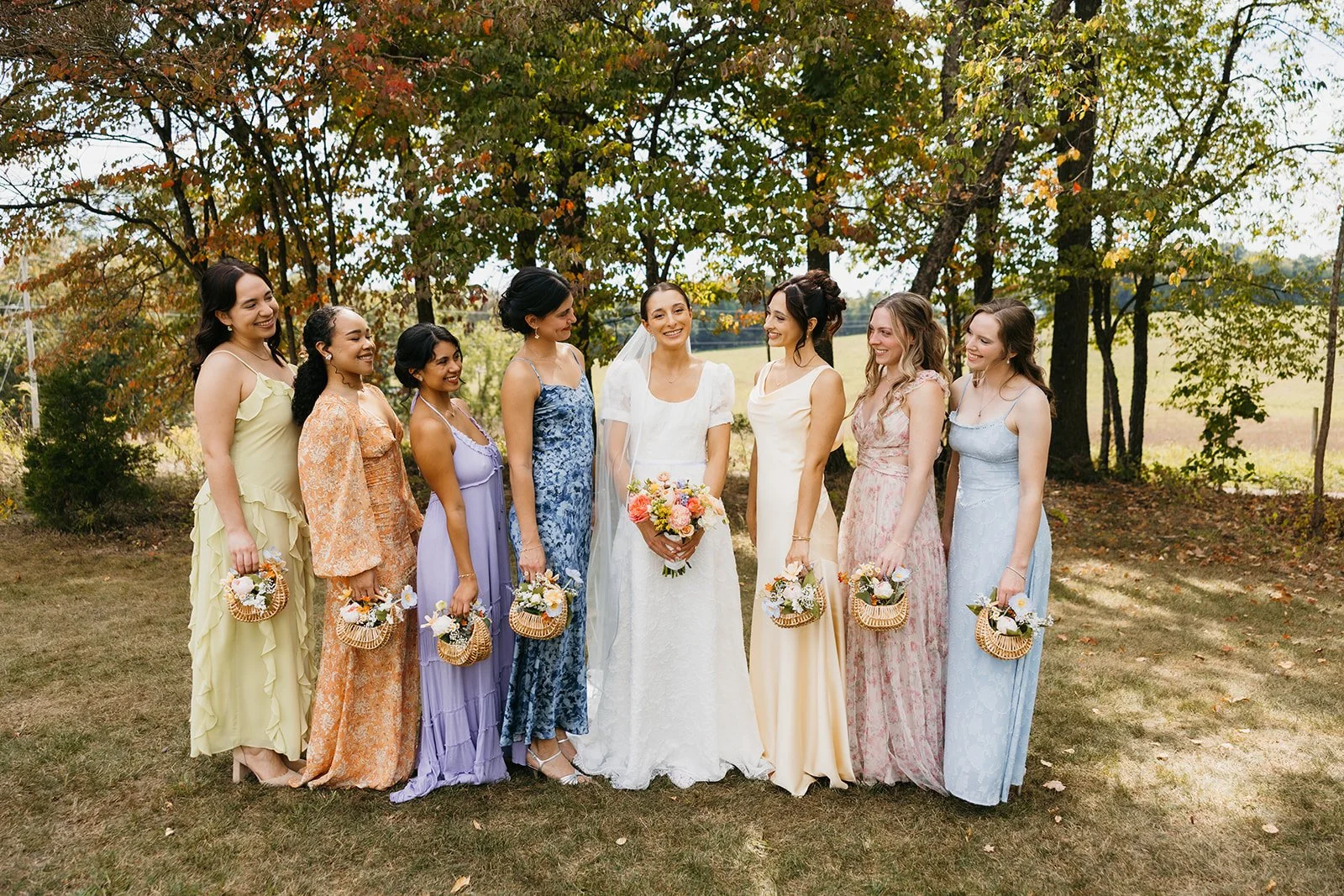 A bride in a white wedding dress holding a bouquet surrounded by eight women in colorful dresses holding wicker baskets, standing outdoors in a grassy area with trees and autumn foliage in the background, smiling and looking at each other.