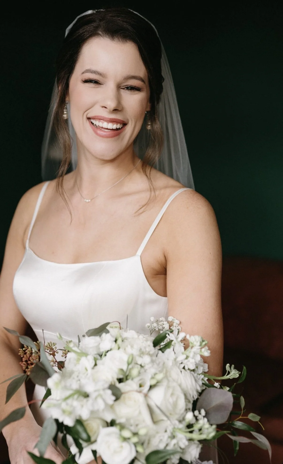 A smiling woman in a white wedding dress with spaghetti straps, holding a bouquet of white flowers, with a sheer veil, dark hair, and earrings, against a dark background.